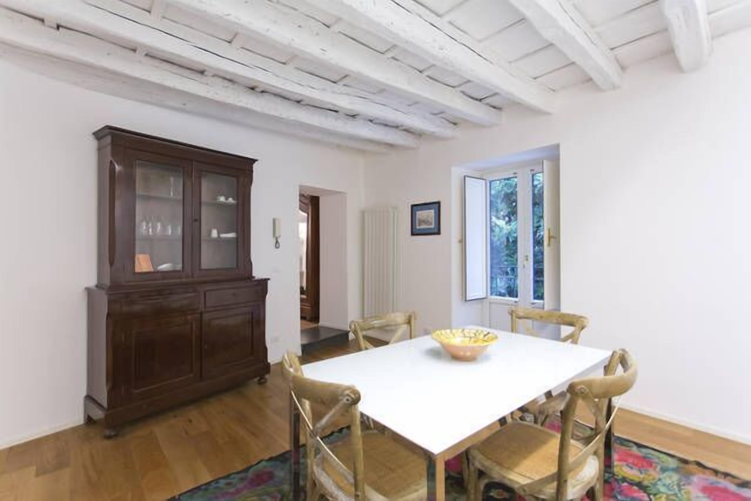 Un área de comedor con una mesa blanca, sillas de madera, un armario de madera oscura y una alfombra colorida.