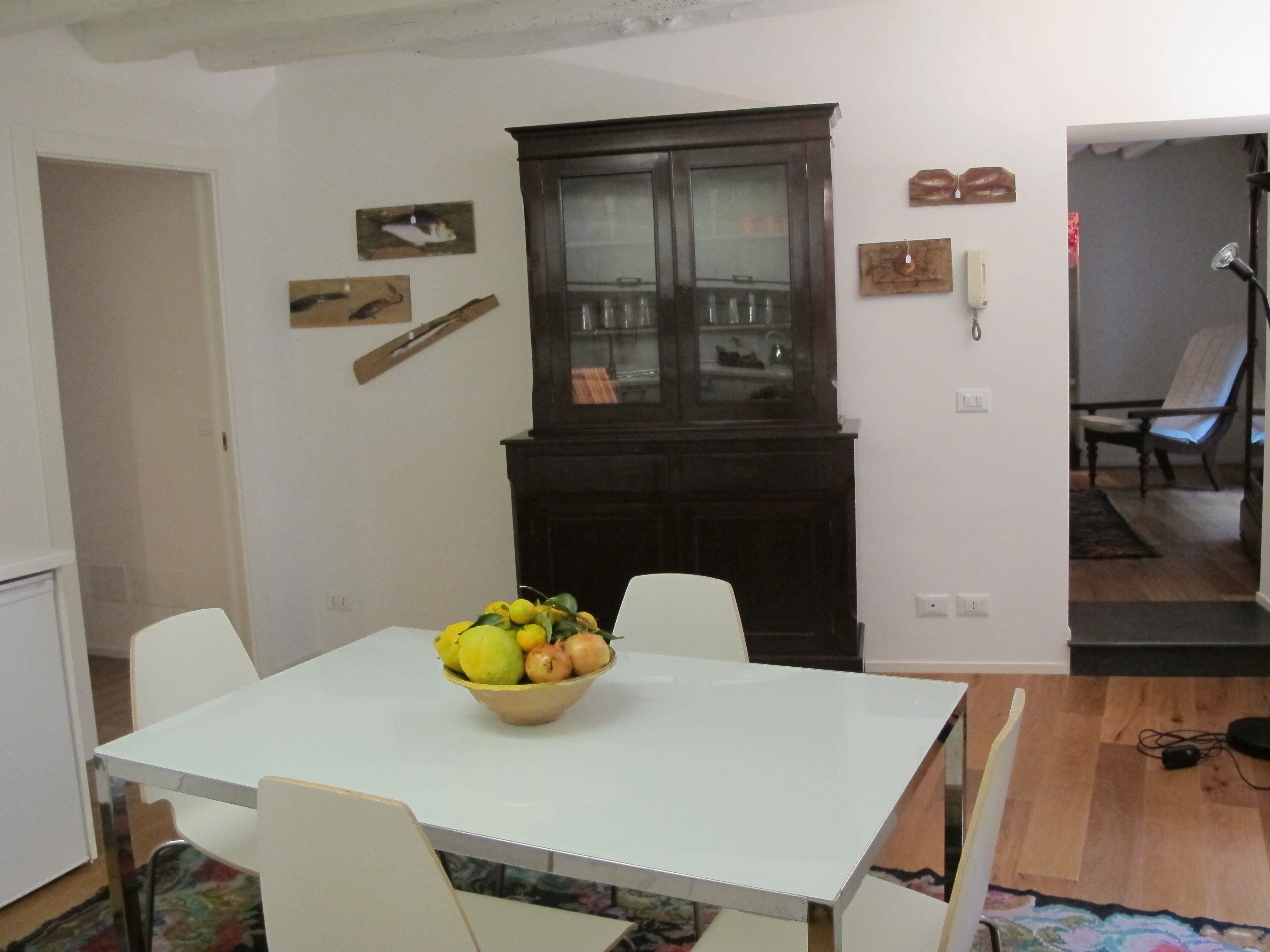 A dining area with a white table, chairs, and a bowl of fruit. There is a dark wooden cabinet with glass doors and a telephone on the wall.