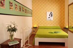 A room with a bed, a bedside table with a book and a vase of flowers, a wall with framed pictures, and a patterned wallpaper. - Prince of Via Veneto DELUXE (Rome)