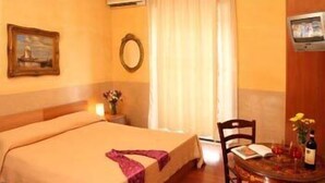 A hotel room with a bed, a small table with a bottle, a chair, a painting, and a TV on the wall. - Prince of Via Veneto DELUXE (Rome)