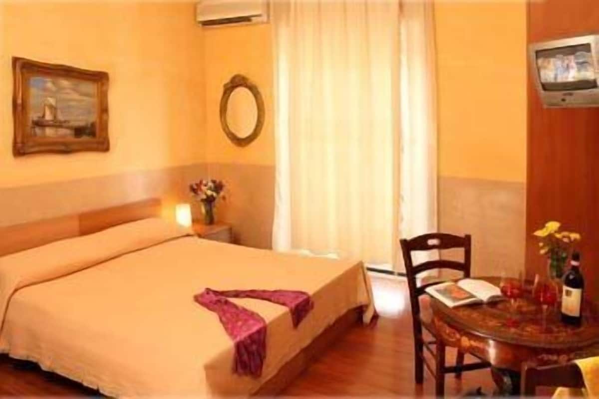 A hotel room with a bed, a small table with a bottle, a chair, a painting, and a TV on the wall.