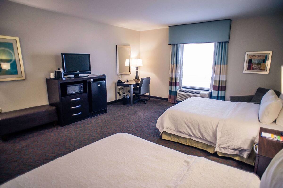 A hotel room with a bed, a desk with a chair, a television, a lamp, and a window with curtains.