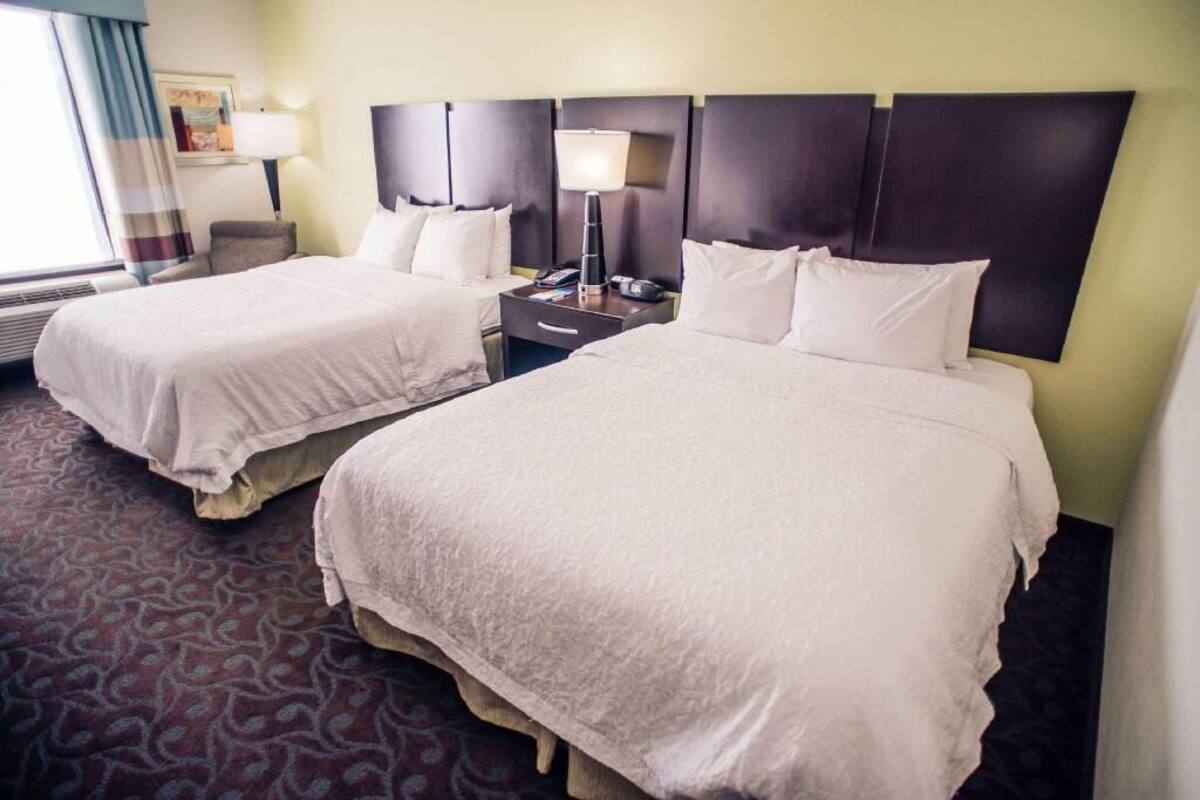 A hotel room with two beds, a nightstand with a lamp, a chair, and a patterned carpet.