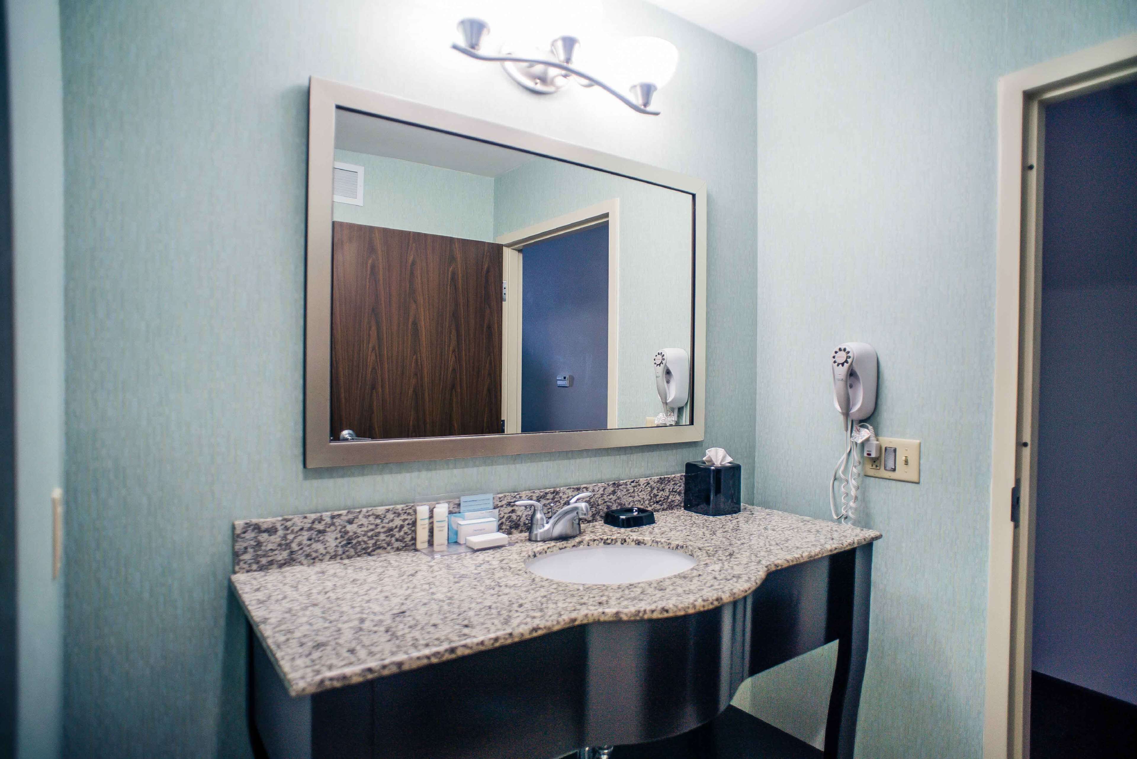 A bathroom with a granite countertop sink, a mirror, a hairdryer, and a towel dispenser.