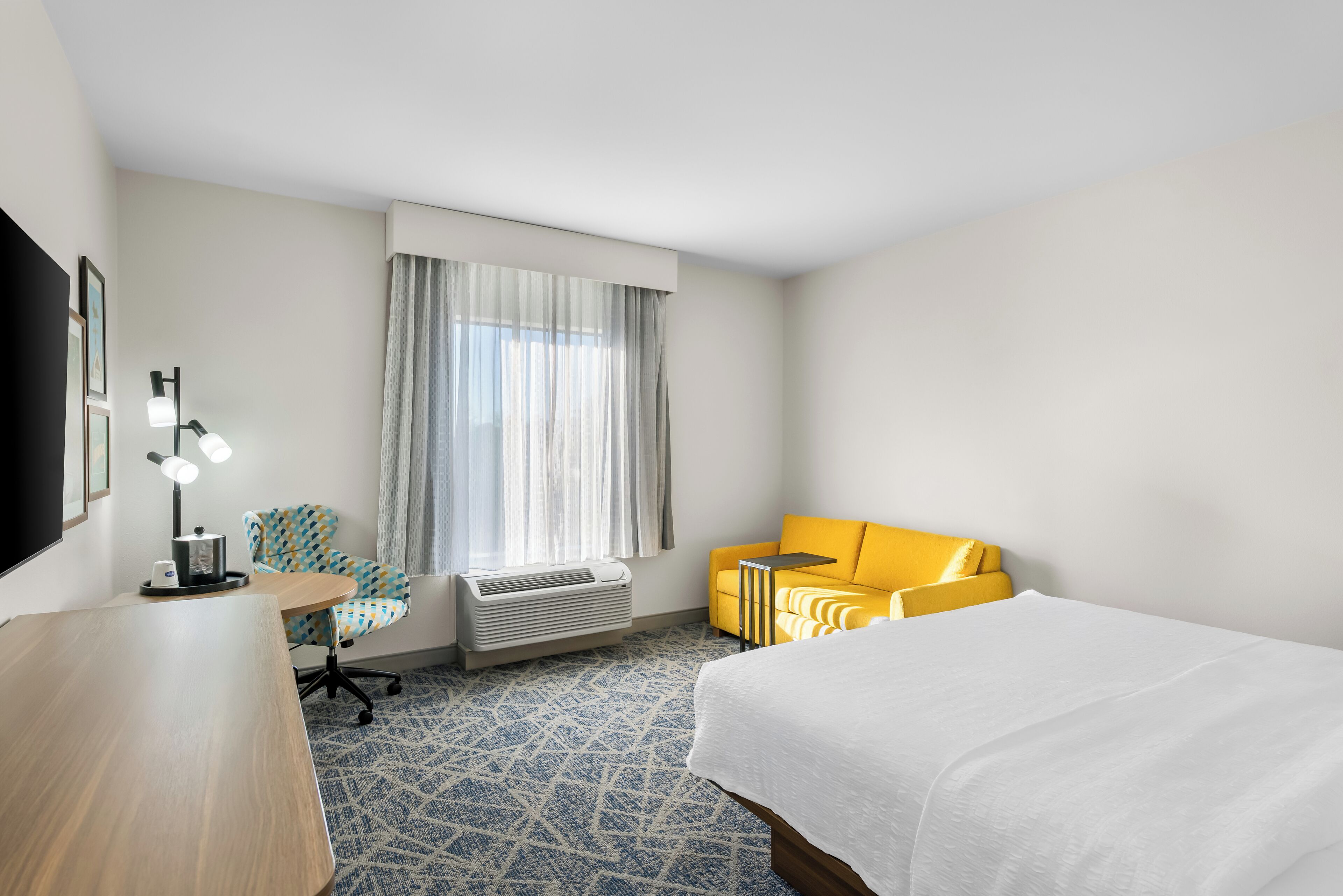 A hotel room with a bed, a desk with a lamp, a chair, a sofa, and a window with curtains.