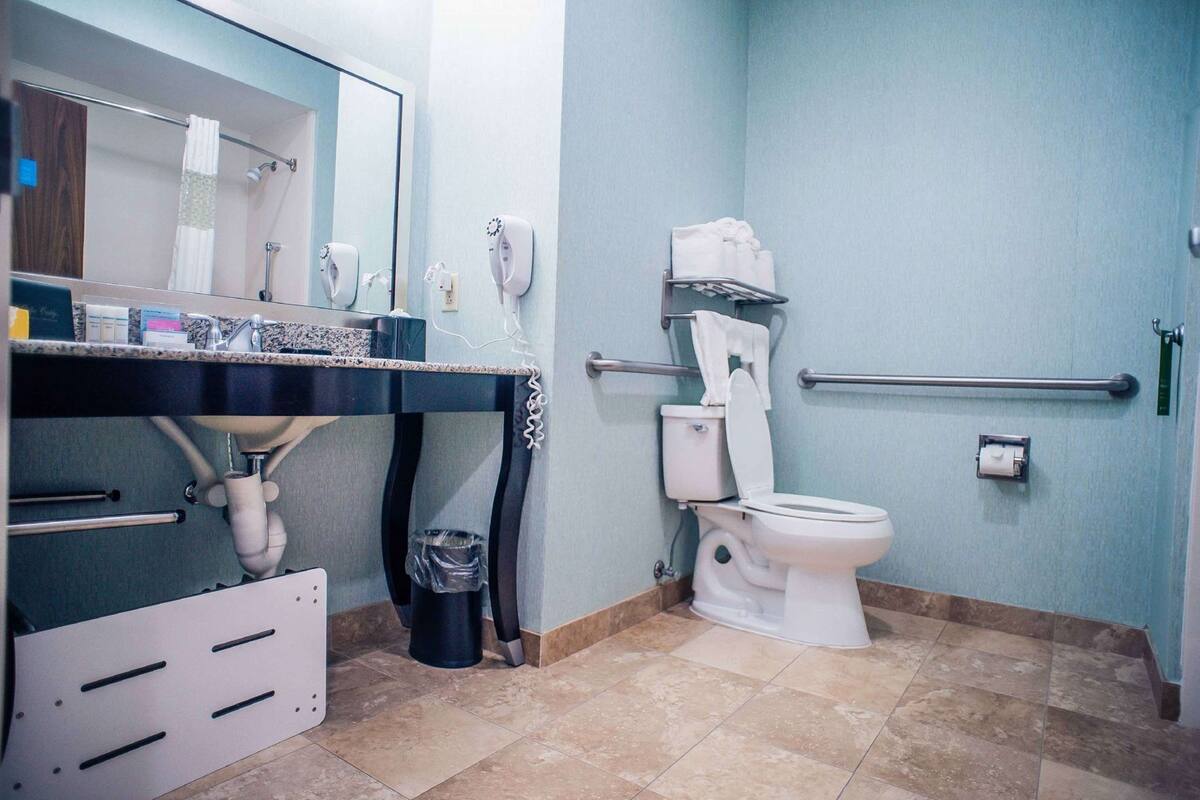 A bathroom with a sink, mirror, toilet, hand dryer, and grab bars.