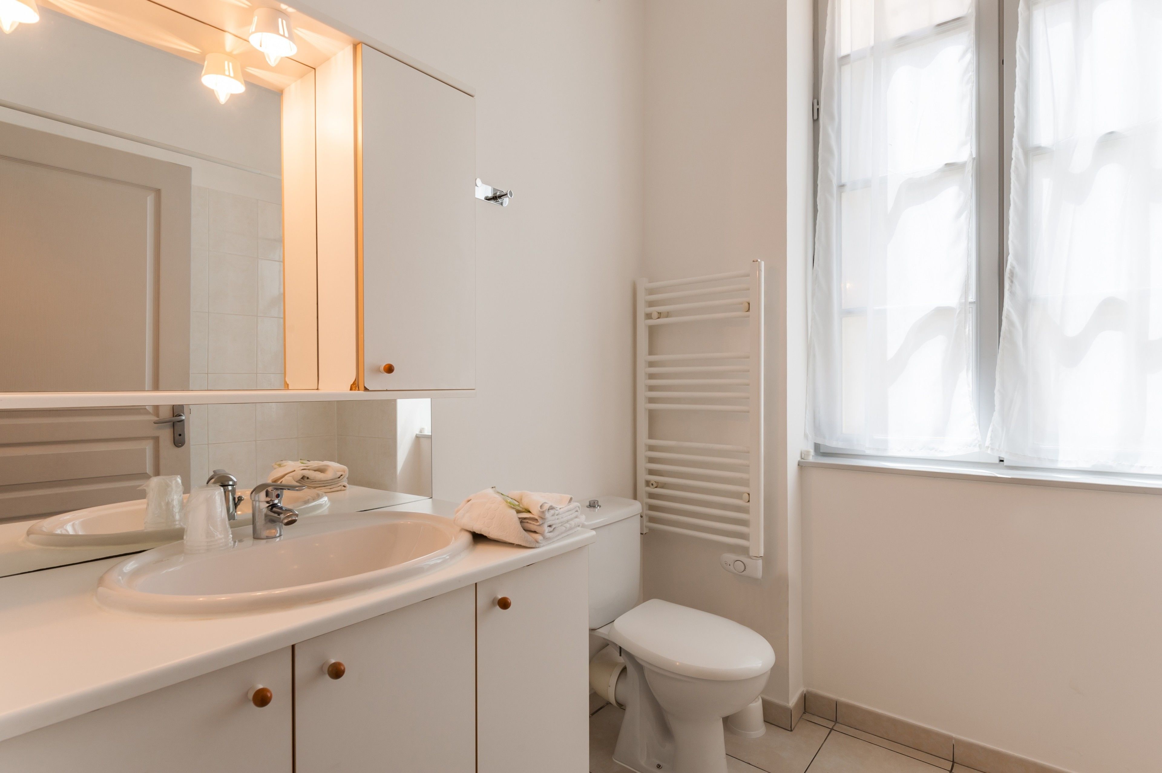 Une salle de bain moderne avec un lavabo blanc, un miroir et des toilettes. On y trouve un porte-serviettes et une fenêtre avec des rideaux légers.