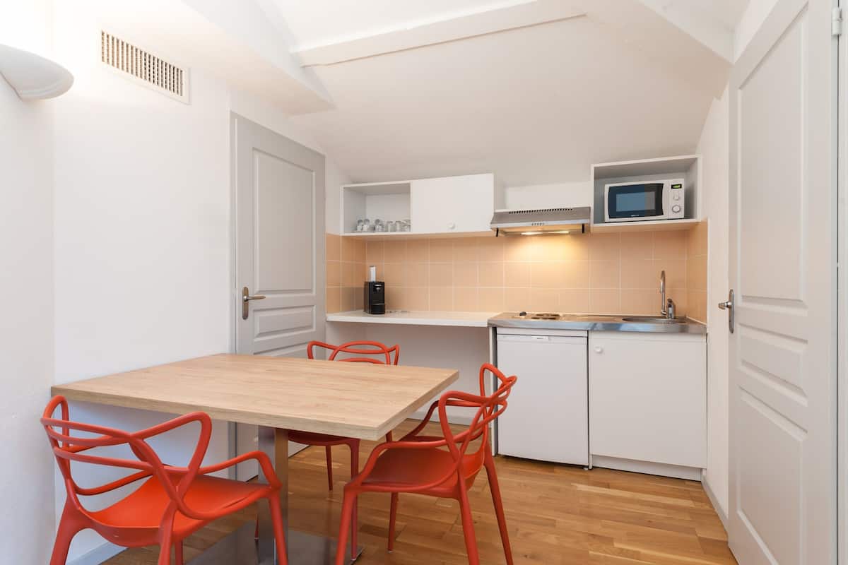 Une cuisine de taille réduite avec des meubles blancs, un four à micro-ondes, un évier et une table en bois accompagnée de chaises rouges.