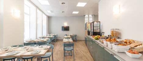 Un comedor luminoso y moderno con mesas largas y sillas azules, una barra de buffet con diversas opciones de comida, y un amplio ventanal que proporciona luz natural.