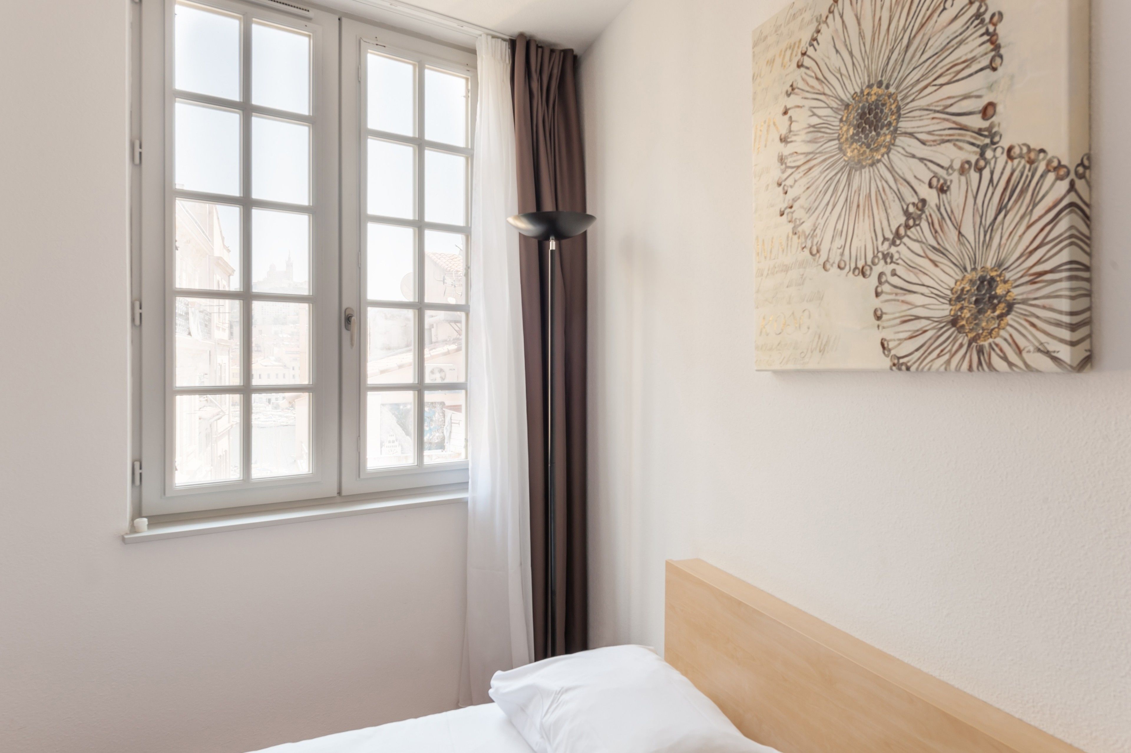 A hotel room with a bed, a window with curtains, and a wall with a floral artwork.