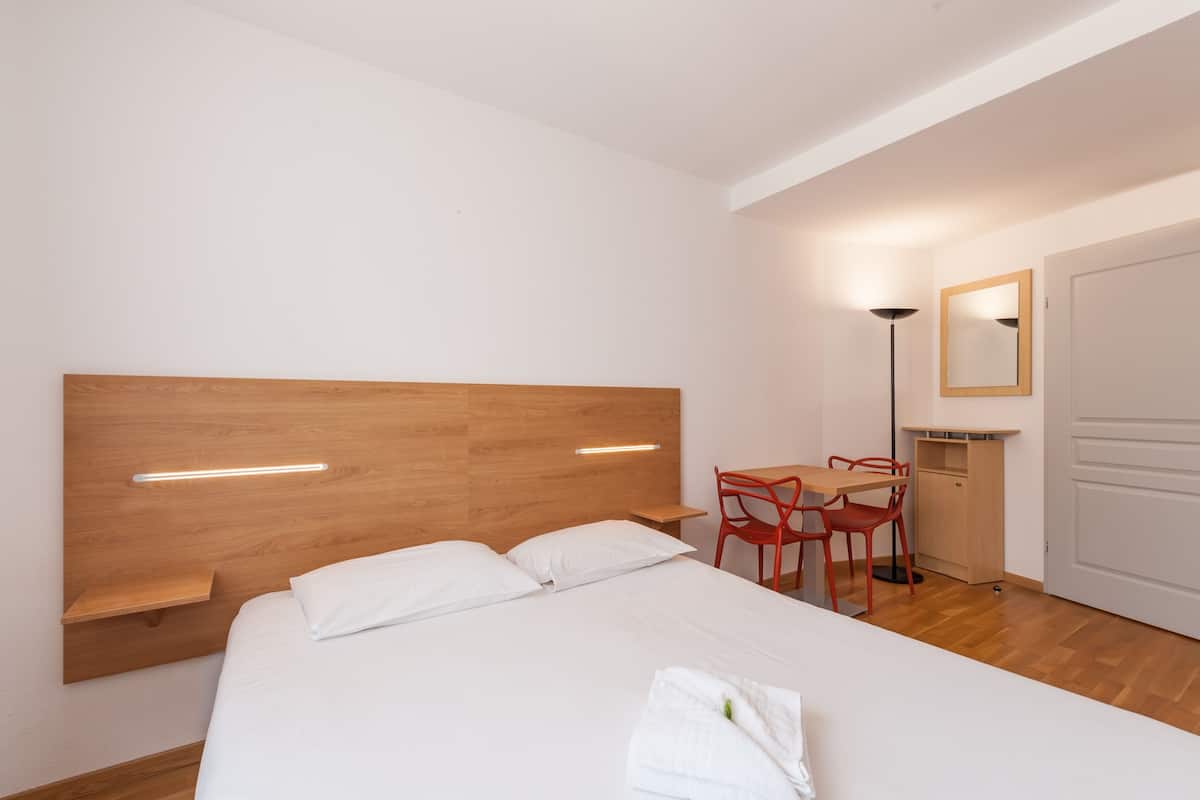 Une chambre d’hôtel avec une tête de lit en bois, un lit avec du linge de maison blanc, un bureau avec une chaise, une lampe sur pied et un miroir au mur.