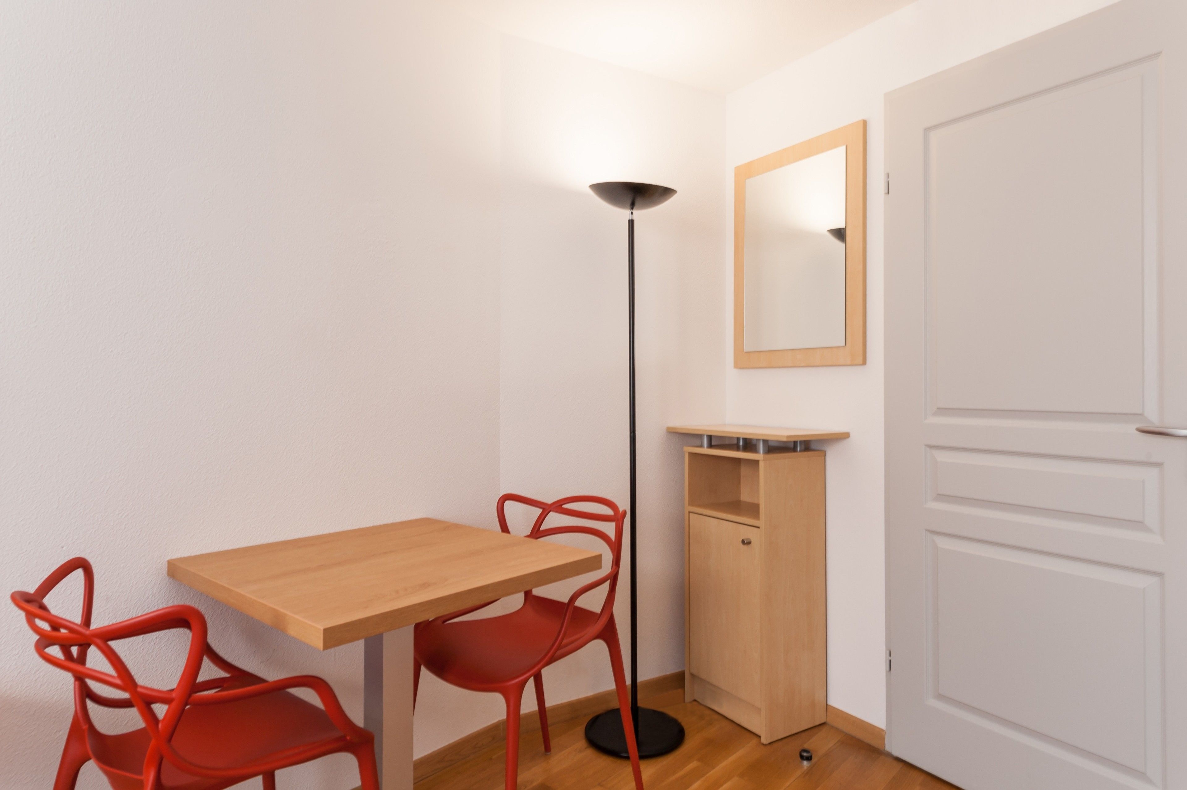 A small room with a wooden table and two red chairs, a floor lamp, a wooden cabinet, and a white door.