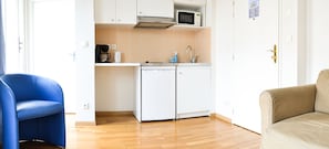 Cocina compacta con microondas, cafetera y lavamanos. Hay una silla azul y un sofá color beige en la sala.