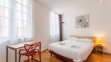 A hotel room with a wooden desk, a red chair, a bed with white linens, a floor lamp, and a wall art piece.