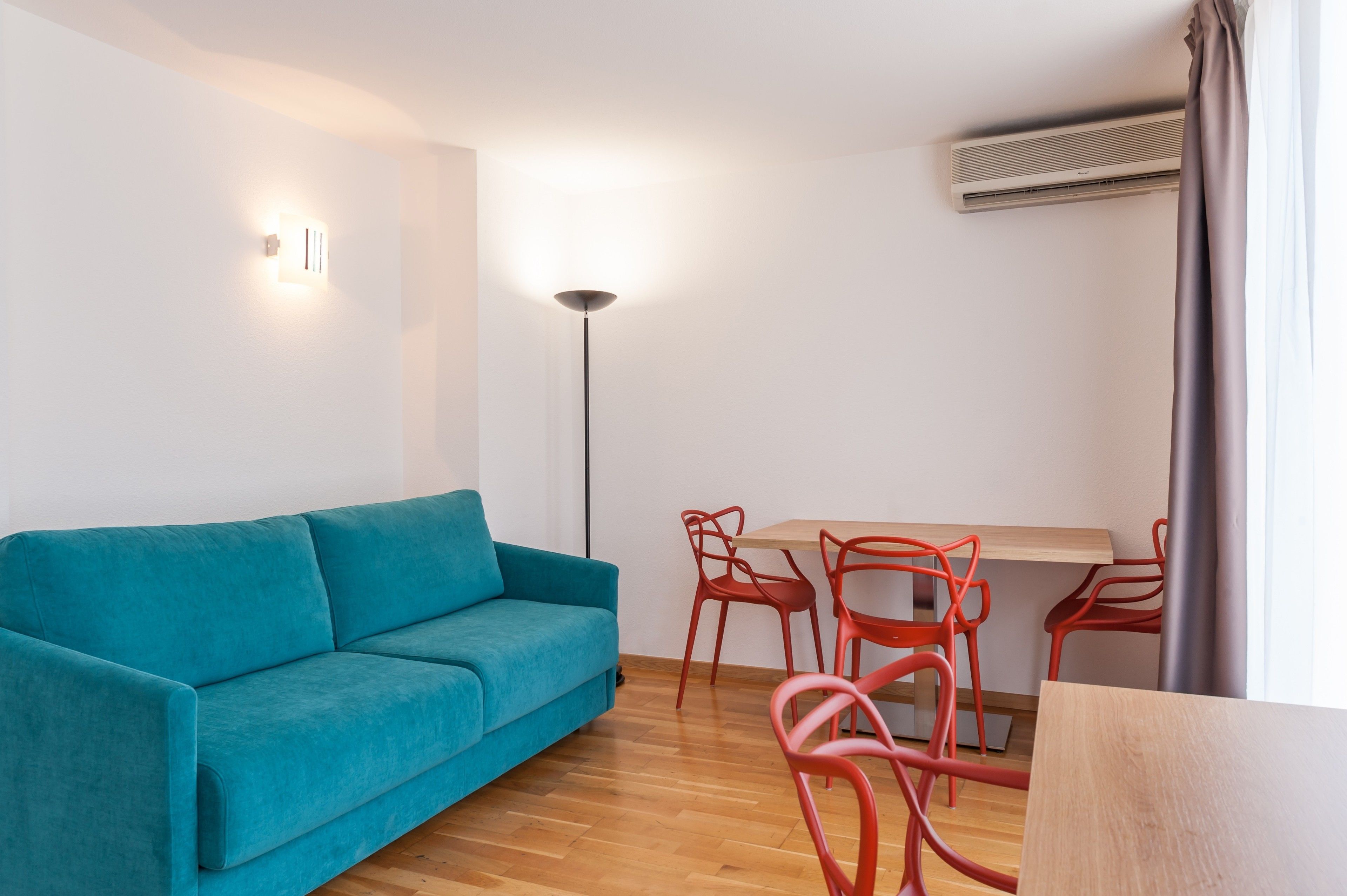 A modern living room with a teal sofa, a wooden dining table with red chairs, a floor lamp, and an air conditioning unit.