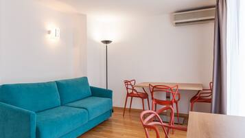A modern living room with a teal sofa, a wooden dining table with red chairs, a floor lamp, and an air conditioning unit.