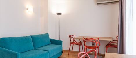 Una sala moderna con un sofá azul verdoso, una mesa de comedor de madera con sillas rojas, una lámpara de pie y un equipo de aire acondicionado.