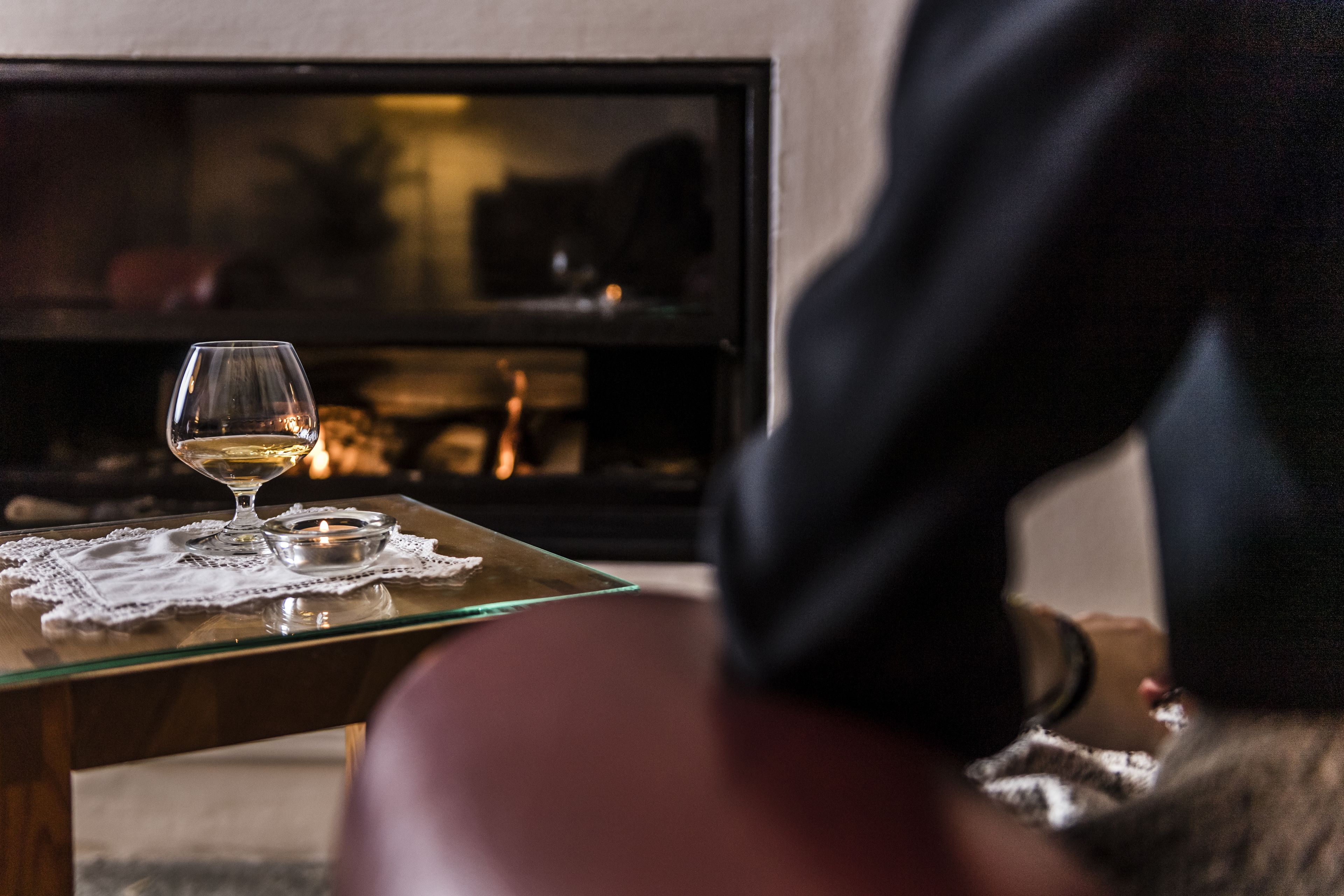 A glass table with a candle and a glass of wine, a fireplace in the background, and a person in the foreground.