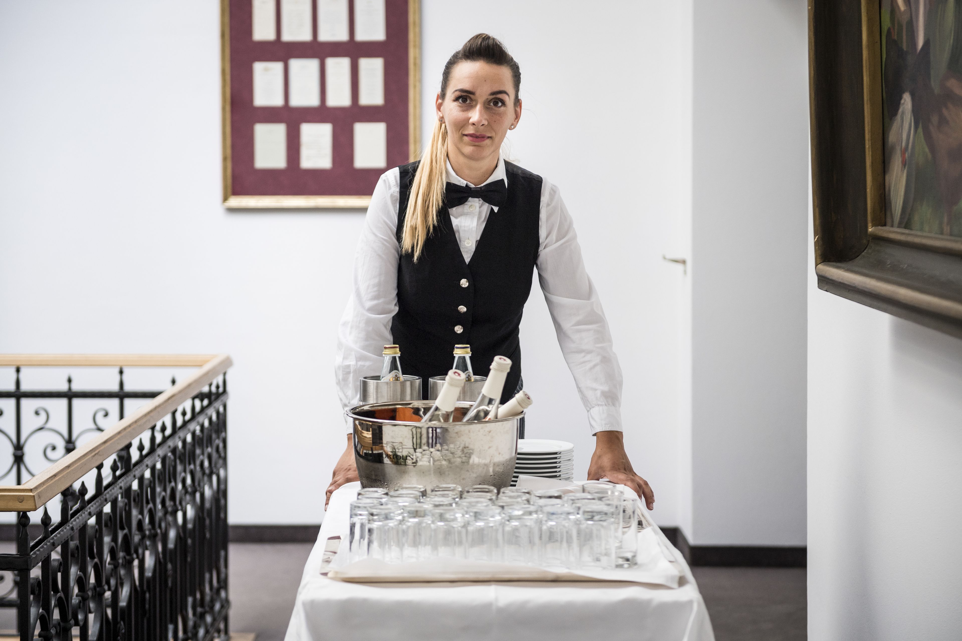 Une personne vêtue d’une chemise blanche et d’un gilet noir, portant un nœud papillon, se tient derrière une table recouverte d’une nappe blanche et tient un seau en argent contenant des bouteilles et des verres.