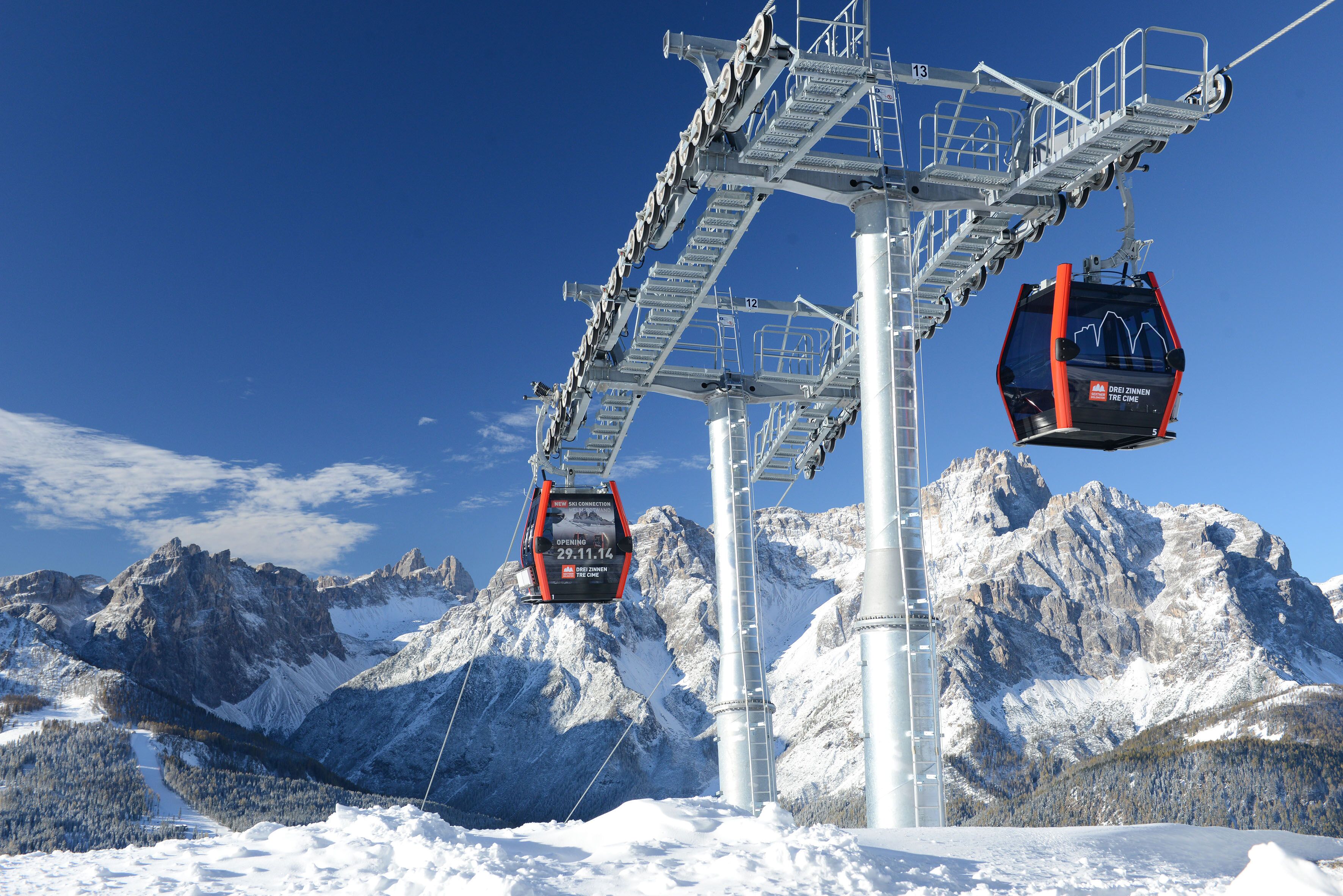 A cable car system with two gondolas suspended from a central tower, set against a backdrop of snow-covered mountains and clear blue sky.