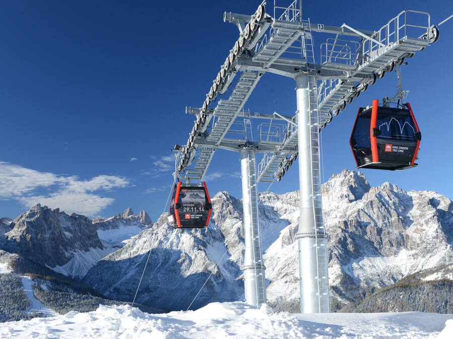 A cable car system with two gondolas suspended from a central tower, set against a backdrop of snow-covered mountains and clear blue sky.