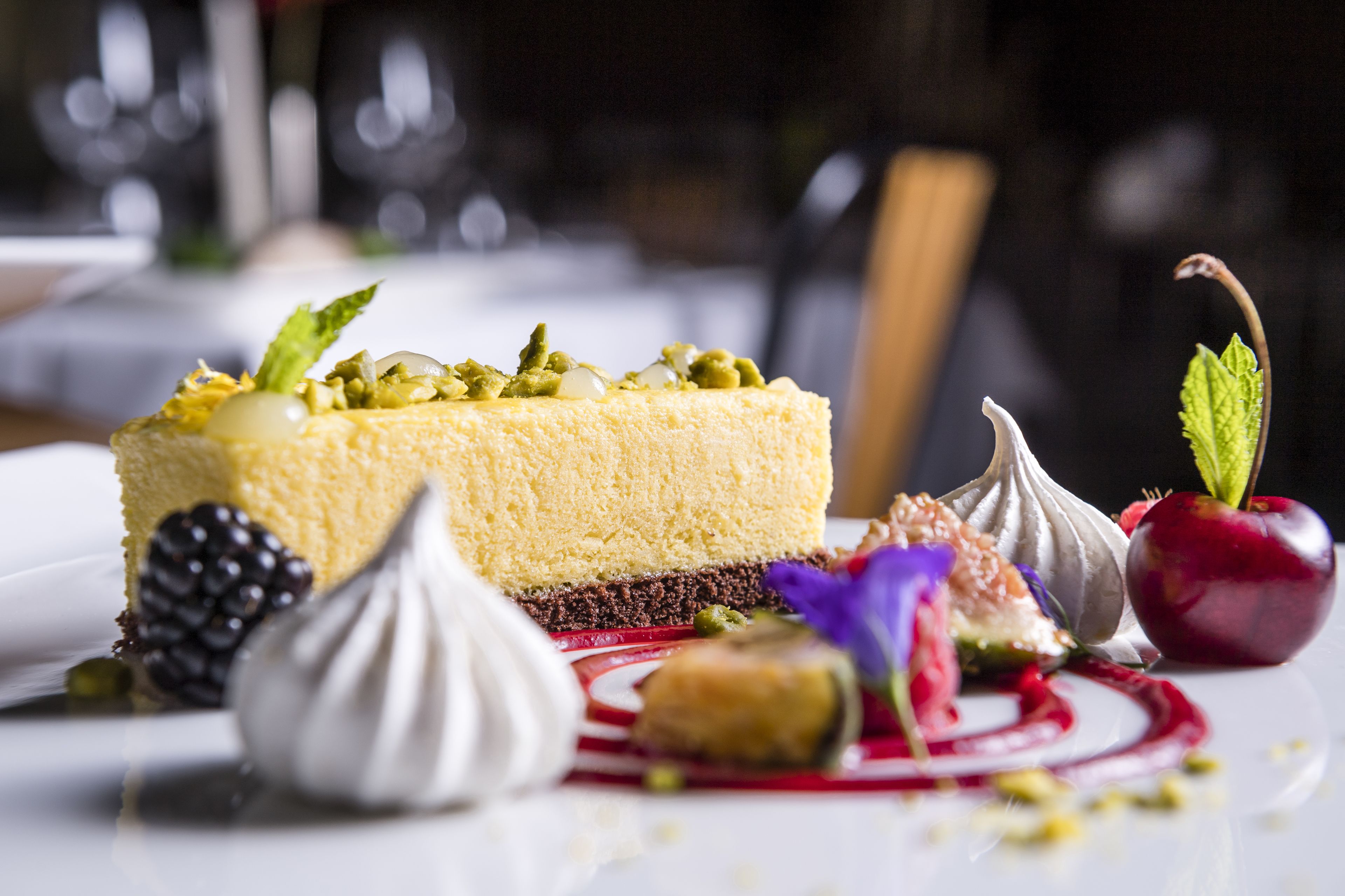 Une assiette à dessert avec une part de gâteau au fromage, décorée de pistaches, d’une mûre et d’une cerise.