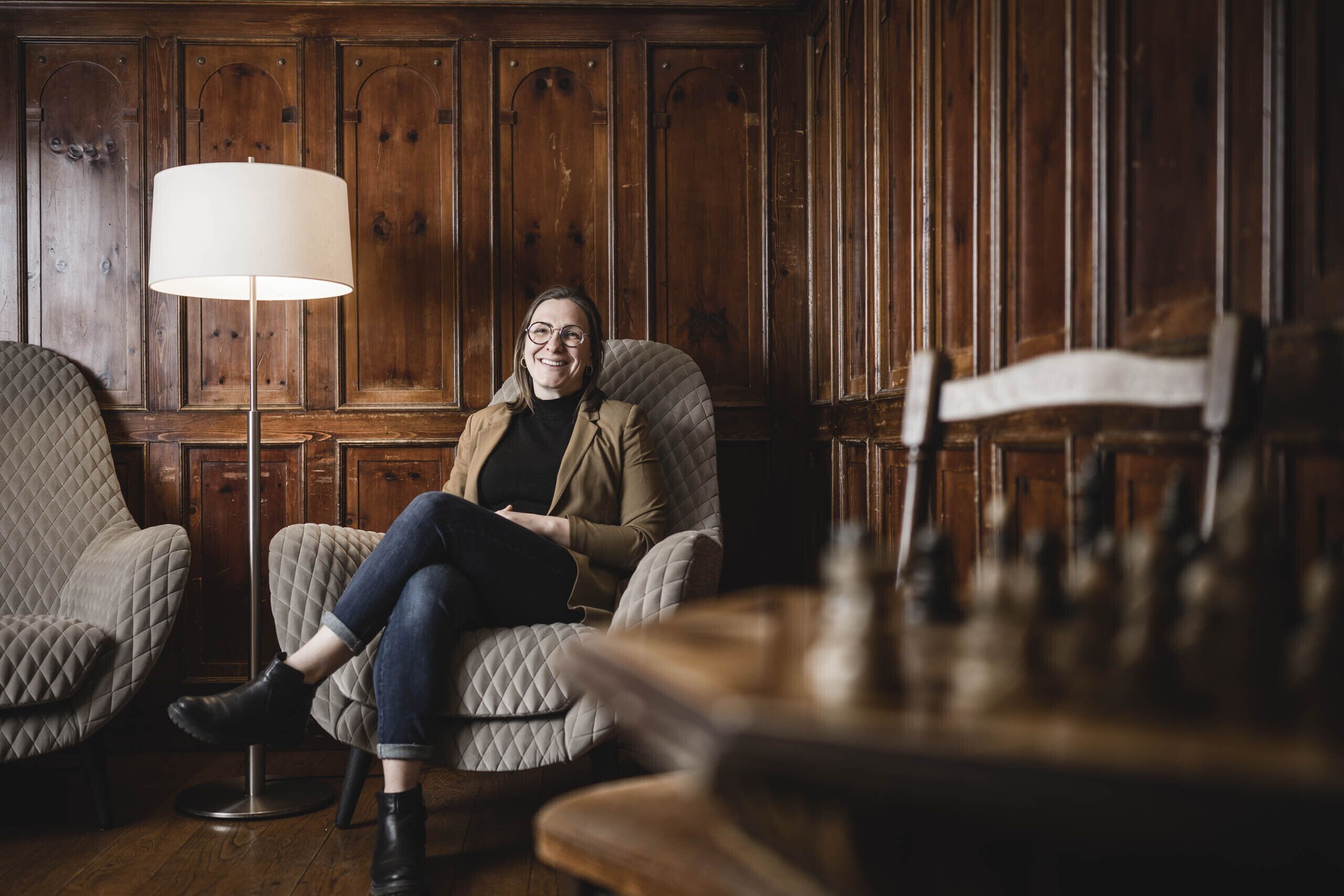 A person seated in a room with wooden paneled walls, a quilted armchair, and a table lamp.