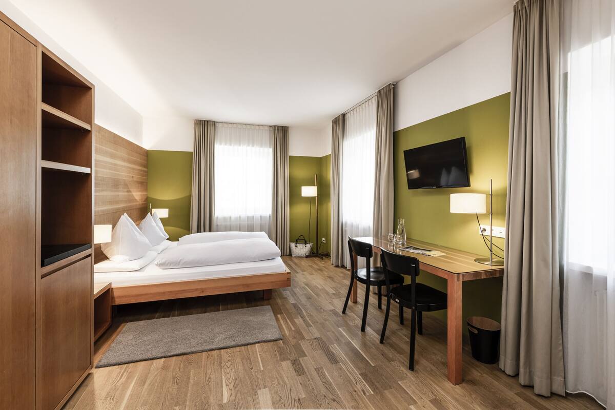 A hotel room with a bed, desk, chair, TV, and a small table with a lamp.