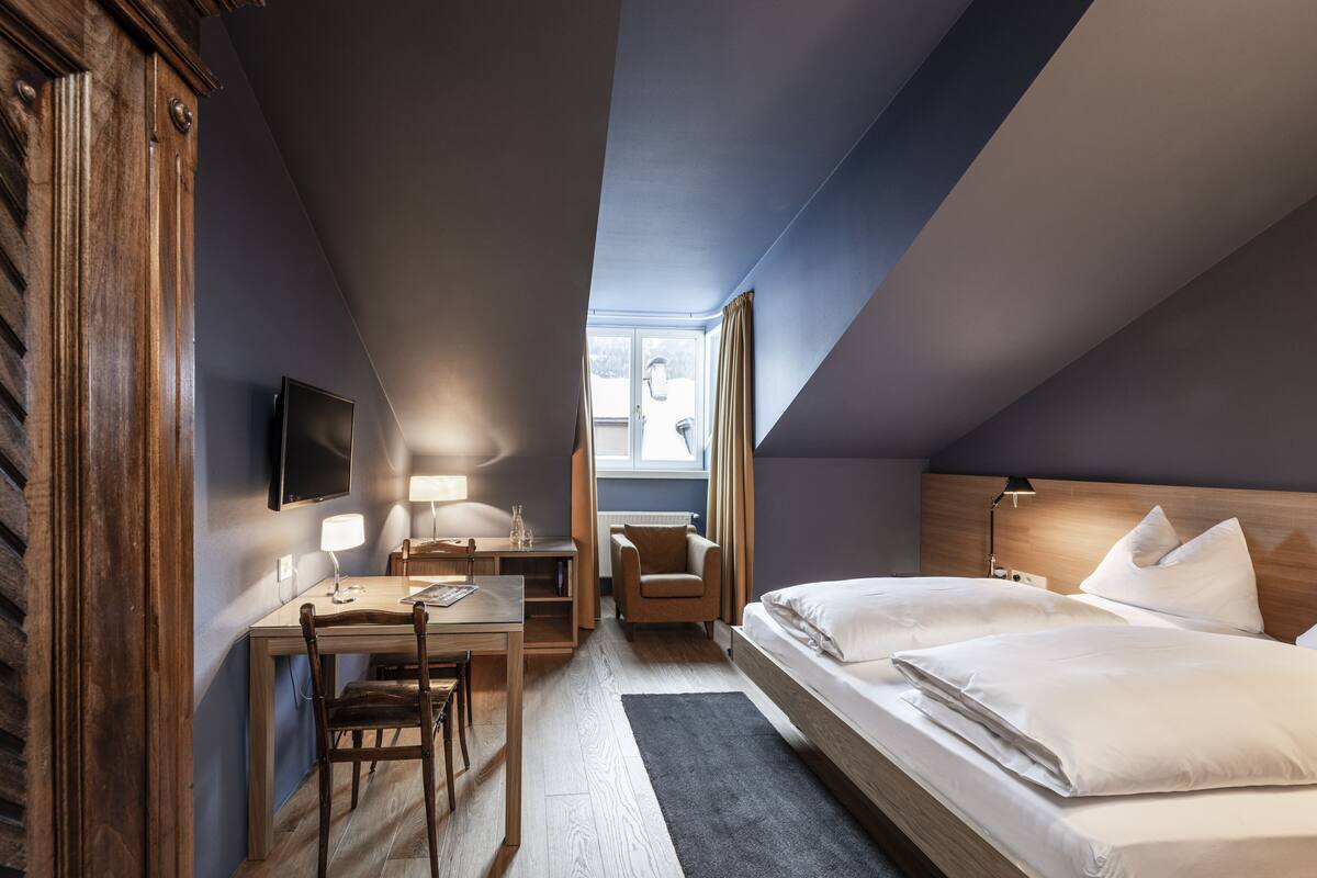 A hotel room with a wooden desk, two beds, a TV, and a window with curtains.