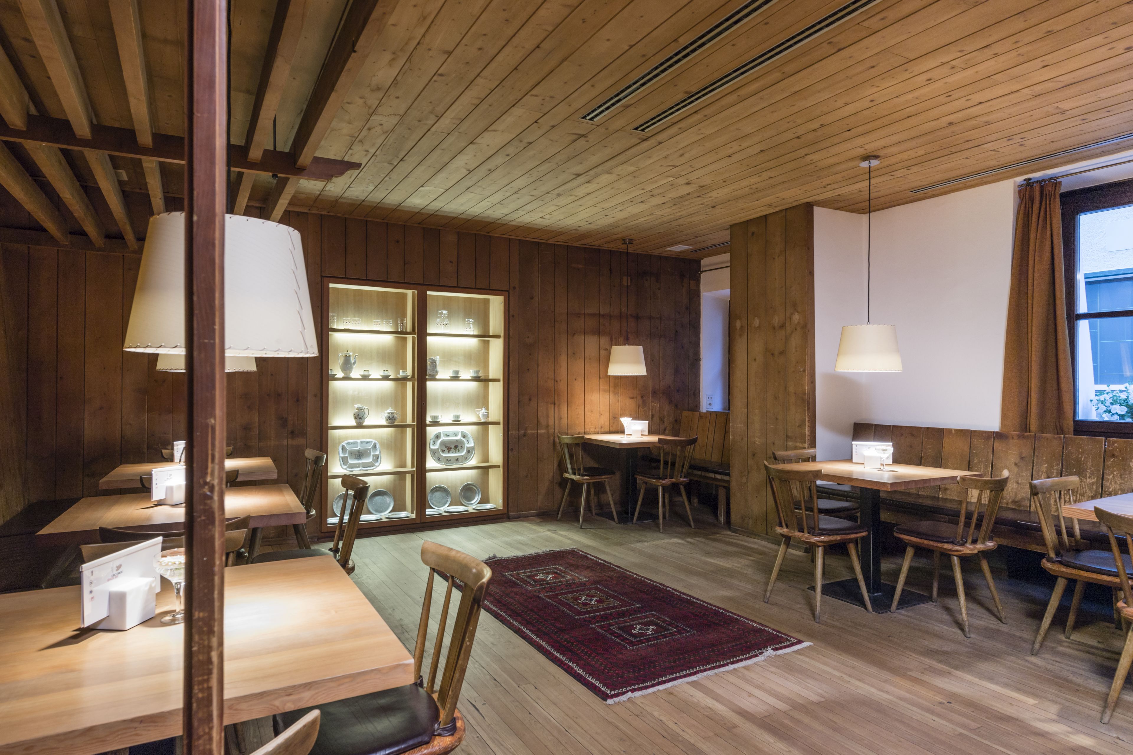 A cozy restaurant with wooden walls and ceiling, featuring tables and chairs, a central display case, and pendant lighting.