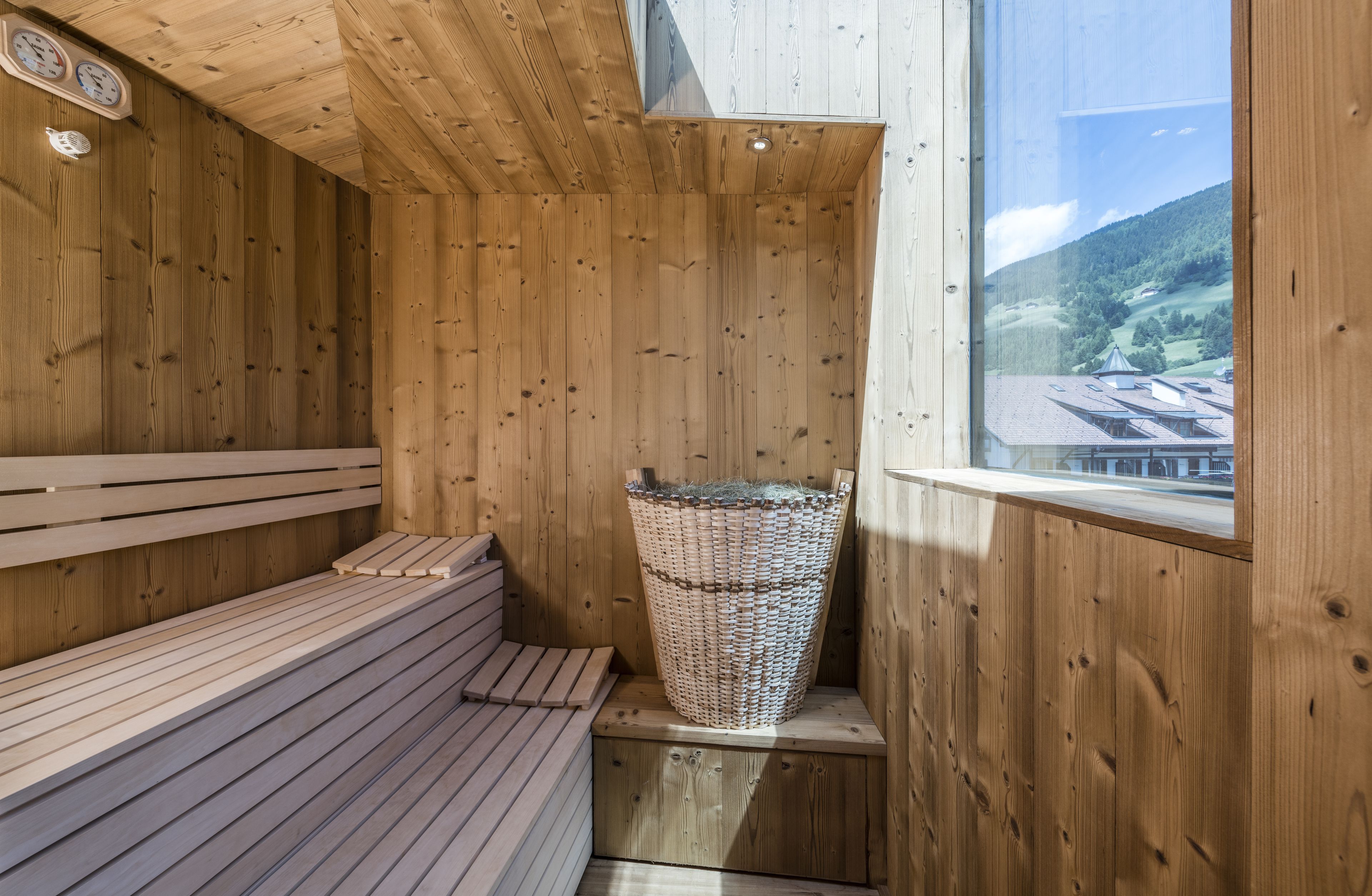 Een houten sauna met een bank, een rieten mand en een raam met uitzicht op gebouwen en heuvels.
