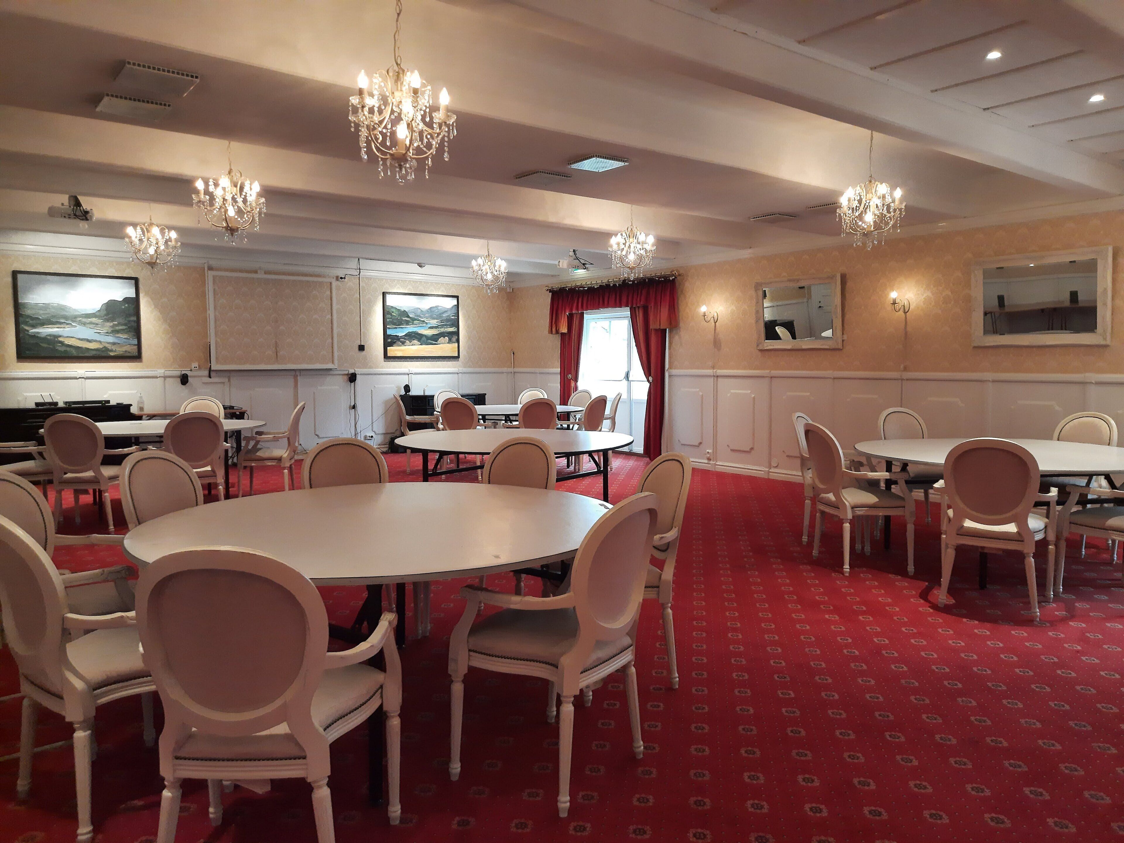 A conference room with round tables and chairs, chandeliers, artwork on the walls, and a red carpeted floor.