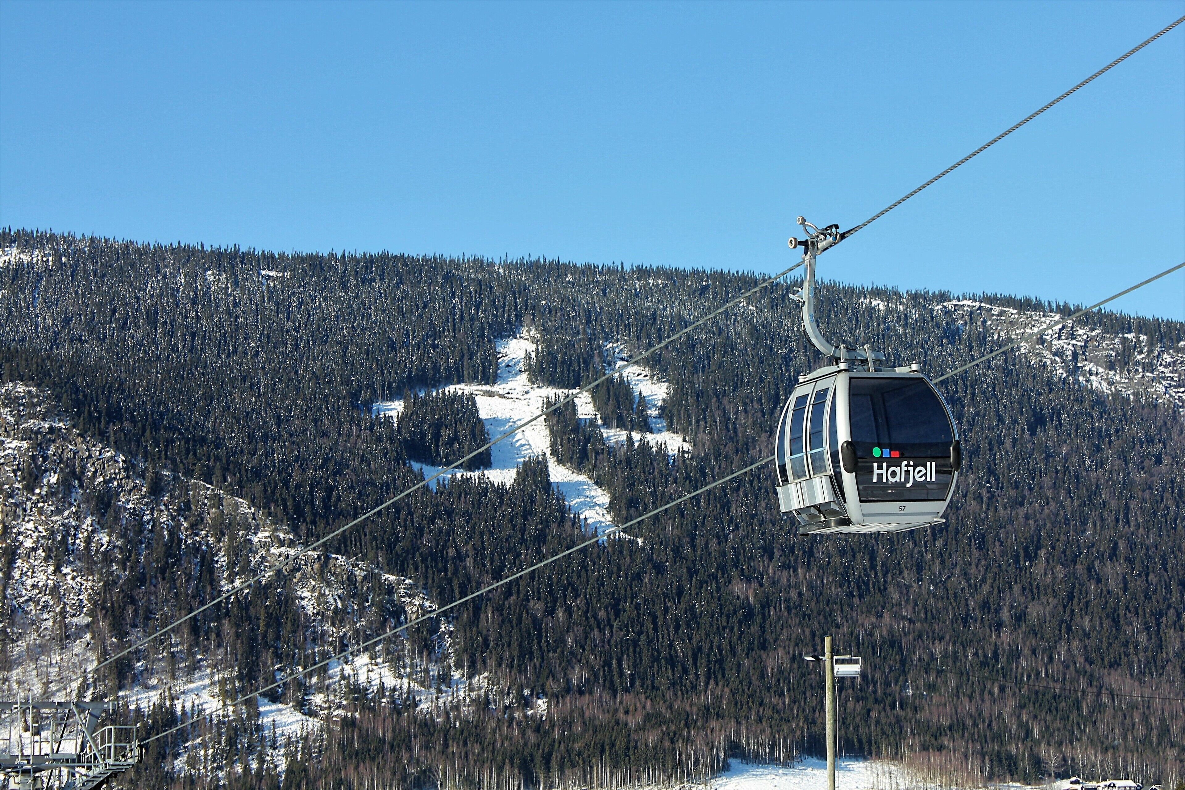 A cable car with the name "Hafjell" on it, moving along a snowy mountain slope with dense forests in the background.