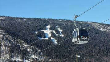 Um teleférico com a inscrição 'Hafjell', movendo-se ao longo de uma encosta nevada de montanha, com densas florestas ao fundo.