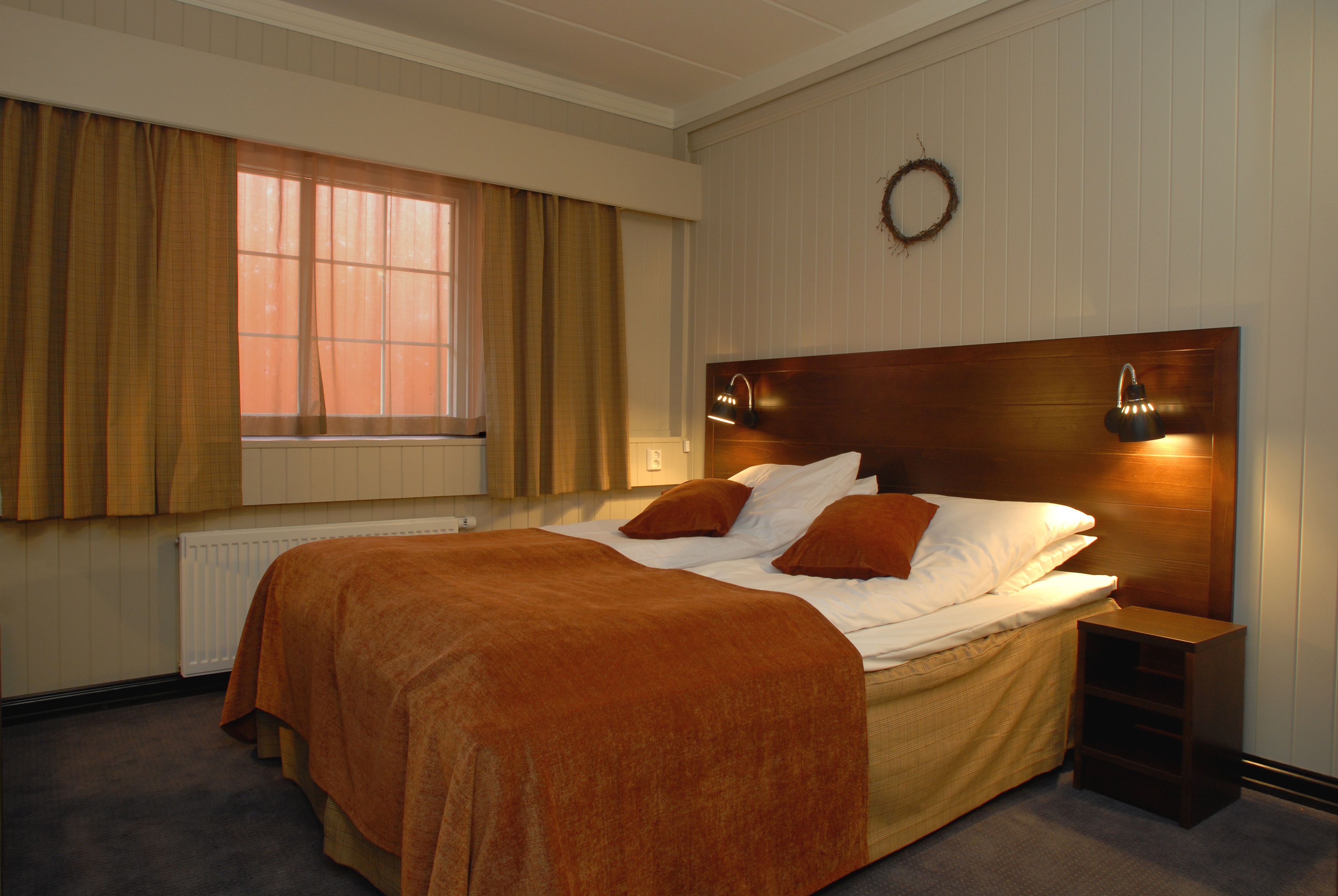 A bedroom with a large bed, a headboard with wall-mounted lights, a window with curtains, and a radiator.