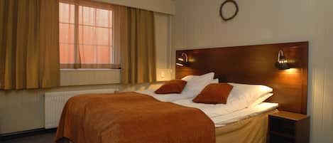 A bedroom with a large bed, a headboard with wall-mounted lights, a window with curtains, and a radiator.