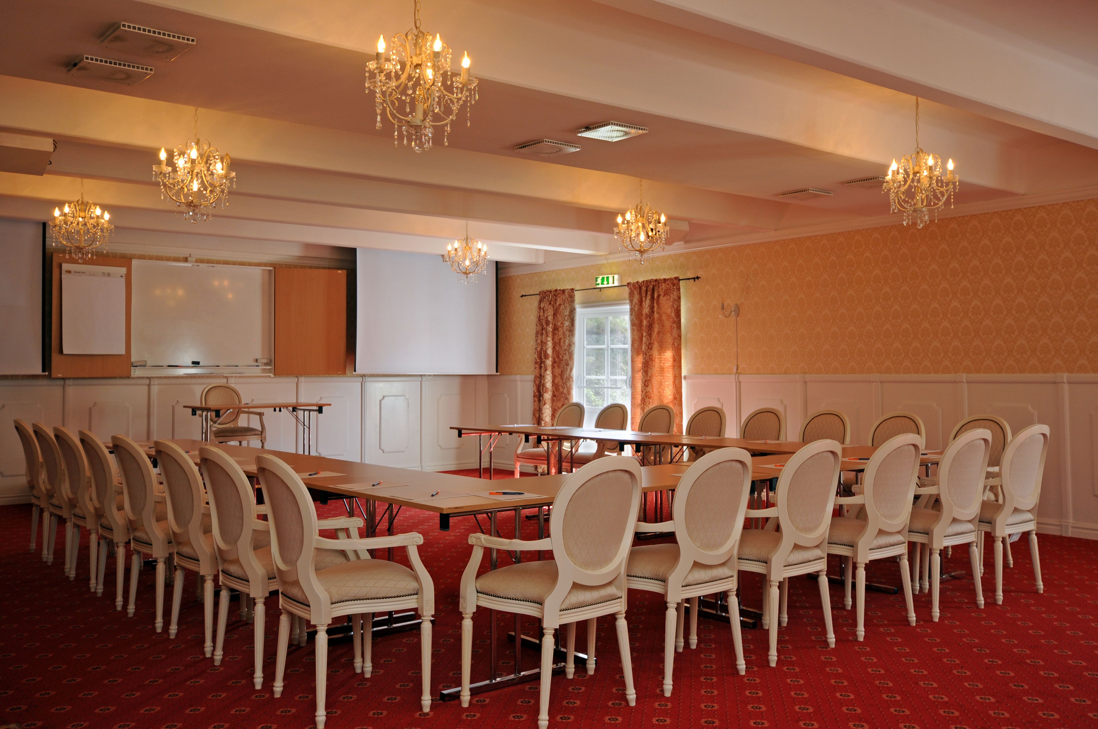 Une salle de conférence avec une longue table, de nombreuses chaises, des lustres, un tableau blanc et un sol recouvert d’une moquette rouge.