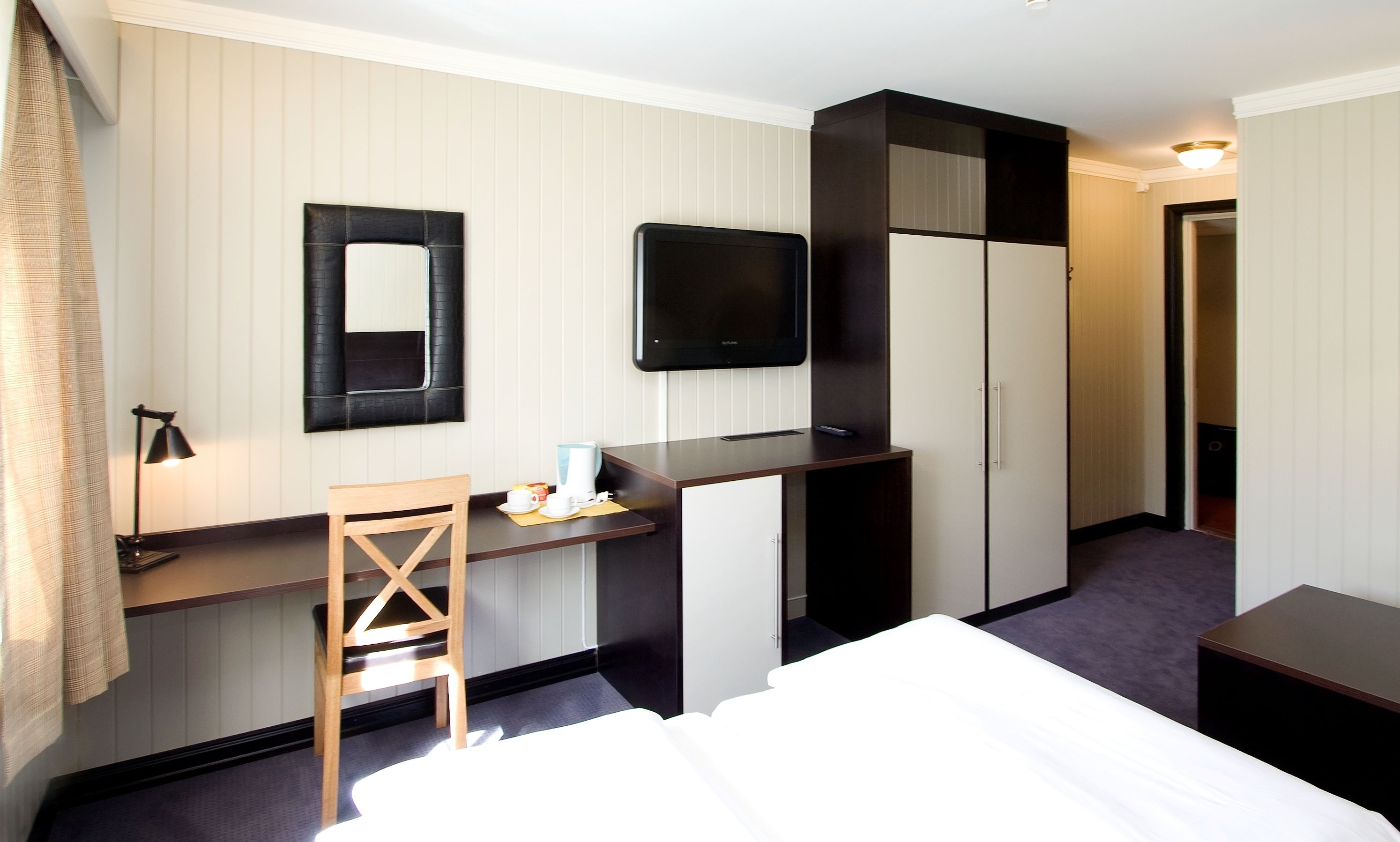 A hotel room with a bed, a desk with a chair, a flat-screen TV, a wardrobe, and a mirror.