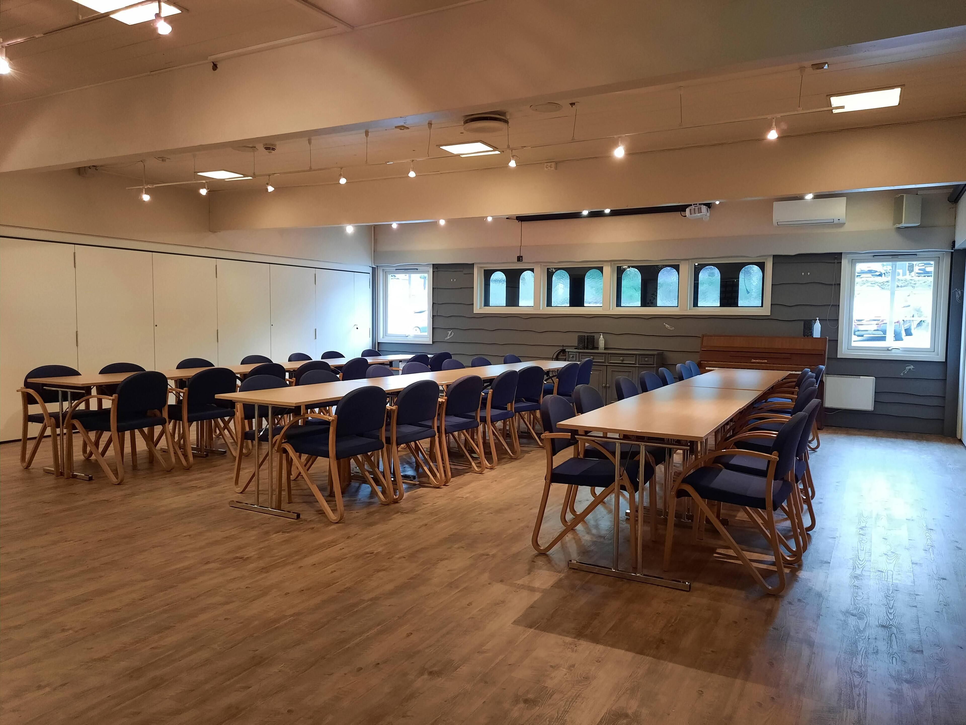 Un salón de conferencias con mesas de madera y sillas azules, ventanas amplias con vidrio esmerilado y luces de techo.