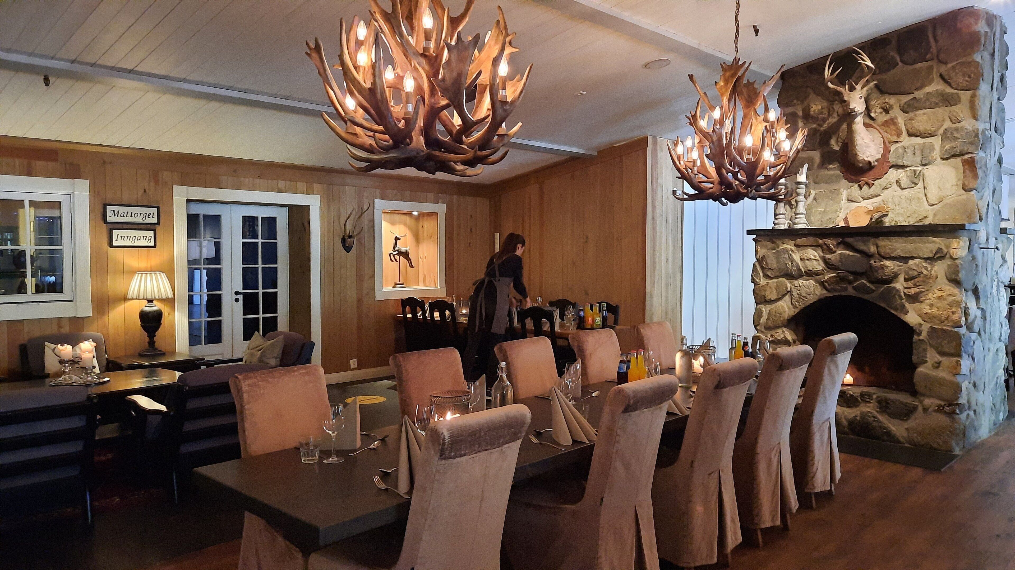 A dining area with a stone fireplace, wooden paneling, and a chandelier with antlers.