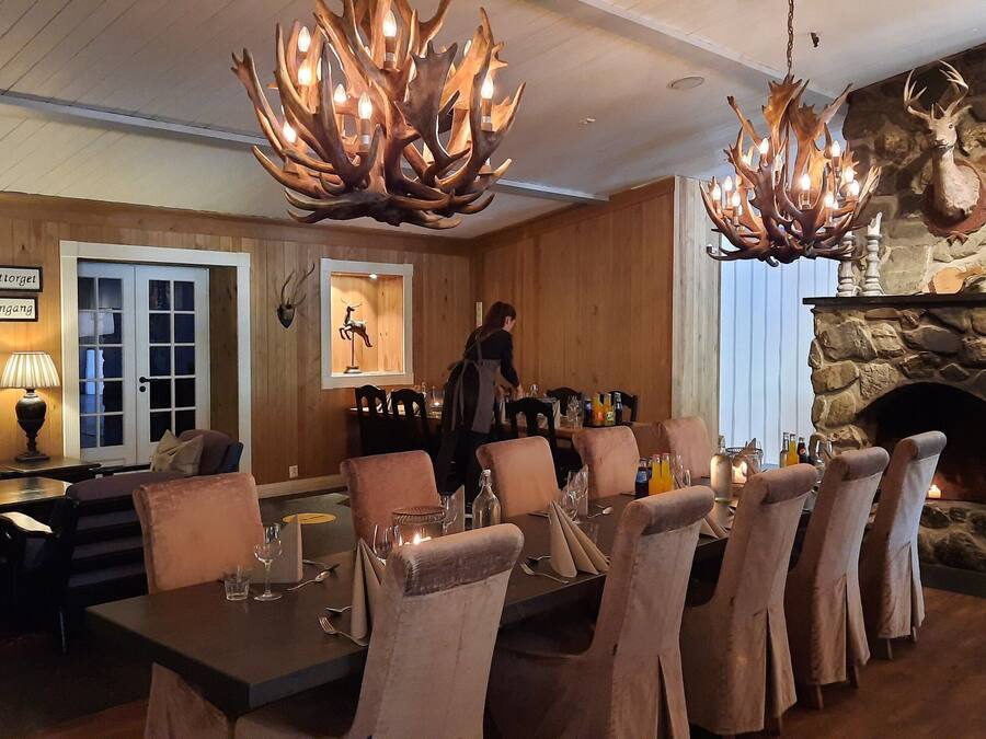 A dining area with a stone fireplace, wooden paneling, and a chandelier with antlers.