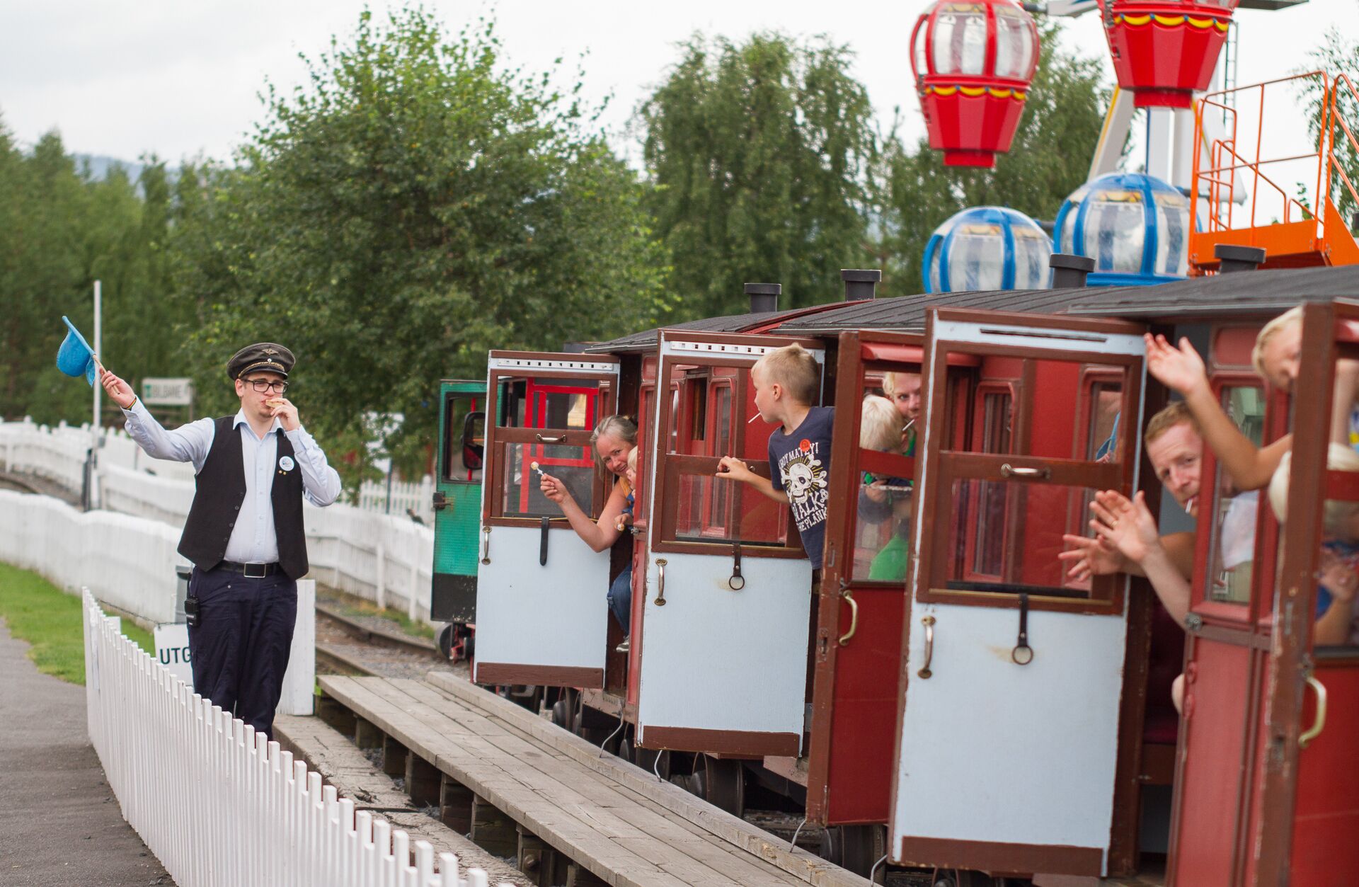 A miniature train with passengers, a conductor, and a track with a white fence.