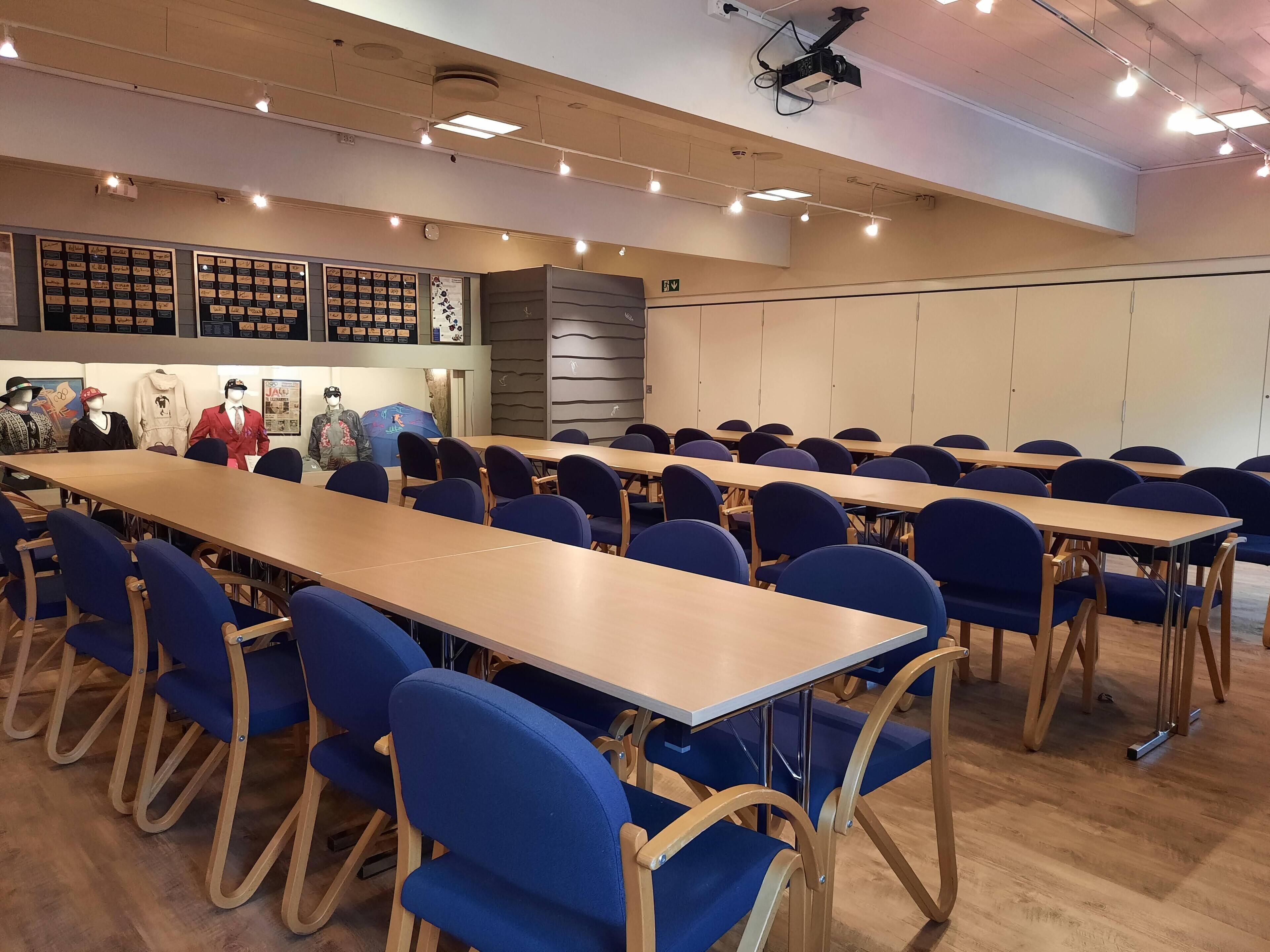 Une salle de conférence avec de longues tables et des chaises bleues, un projecteur au plafond et un mur orné de différents objets encadrés.