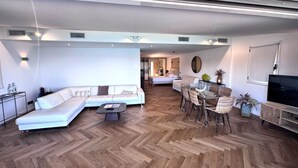 A modern living room with a sofa, dining table, and chairs, a TV, and a hallway leading to a bedroom. - Escale Côté Sud (L'Île-Rousse)