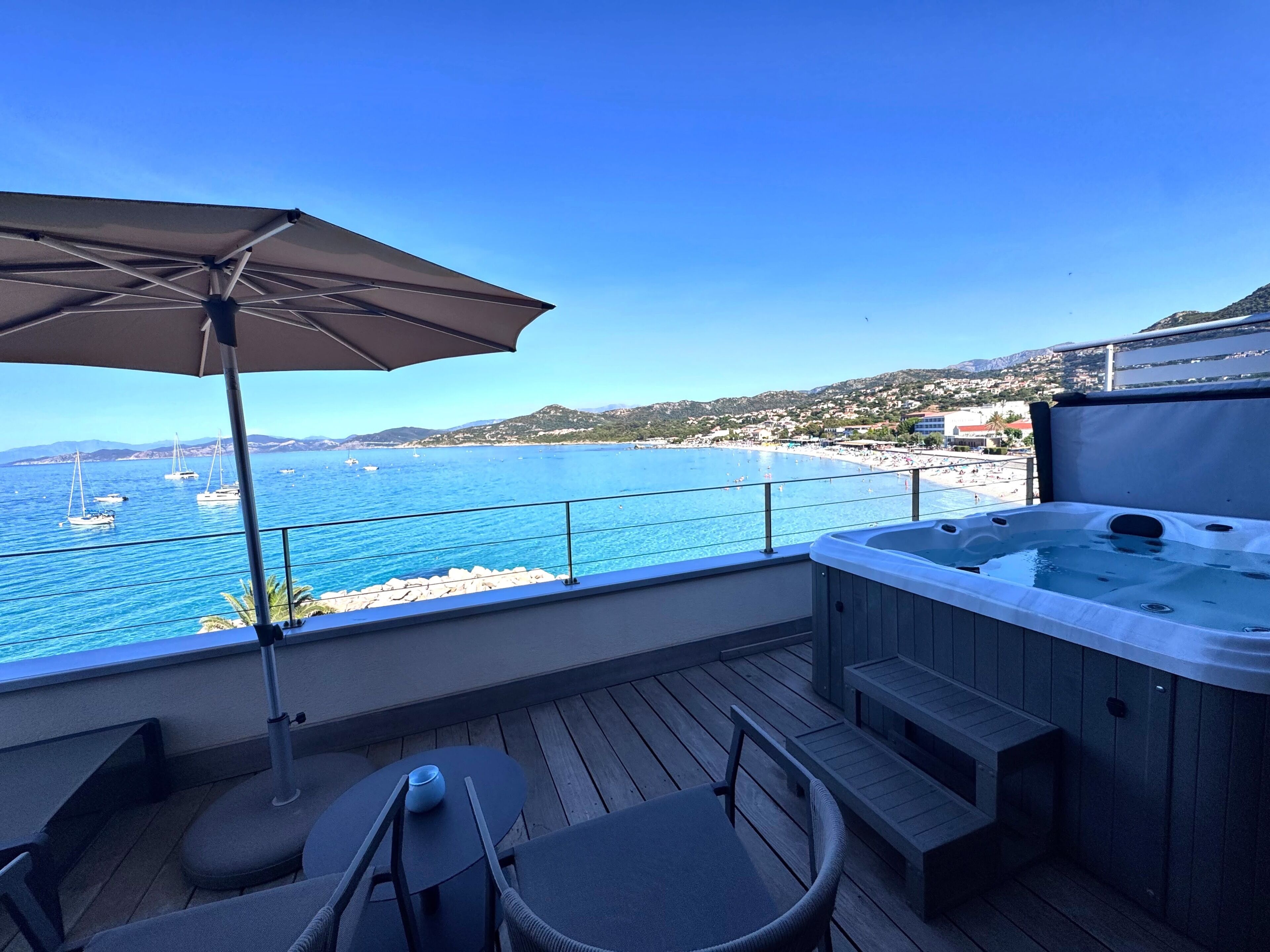 Une terrasse en bois avec un jacuzzi, un espace salon extérieur et une vue sur la mer et la plage.