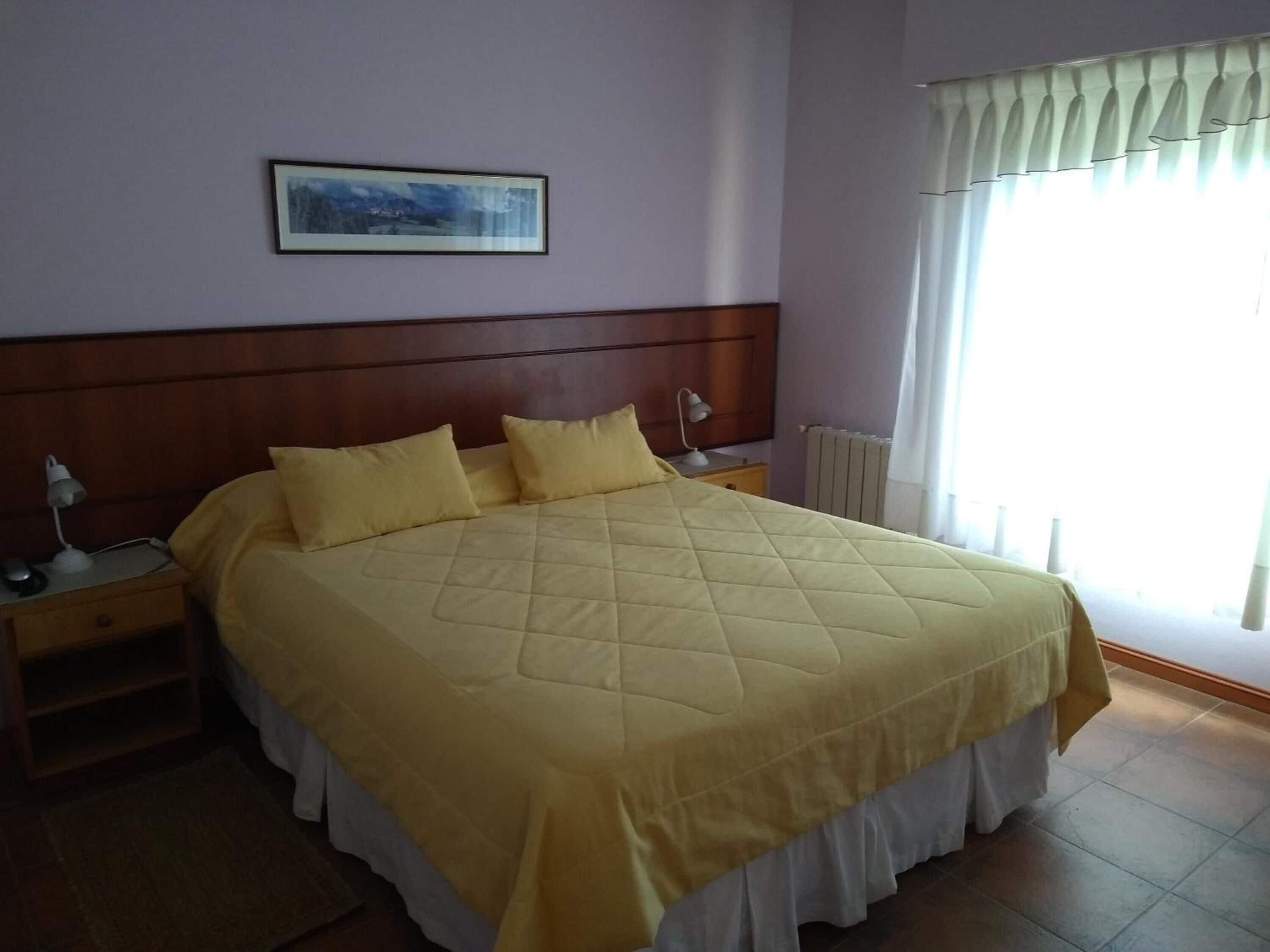 Ein Schlafzimmer mit einem großen Bett, einem Nachttisch, einer Lampe, einem gerahmten Bild an der Wand und einem Fenster mit Vorhängen.