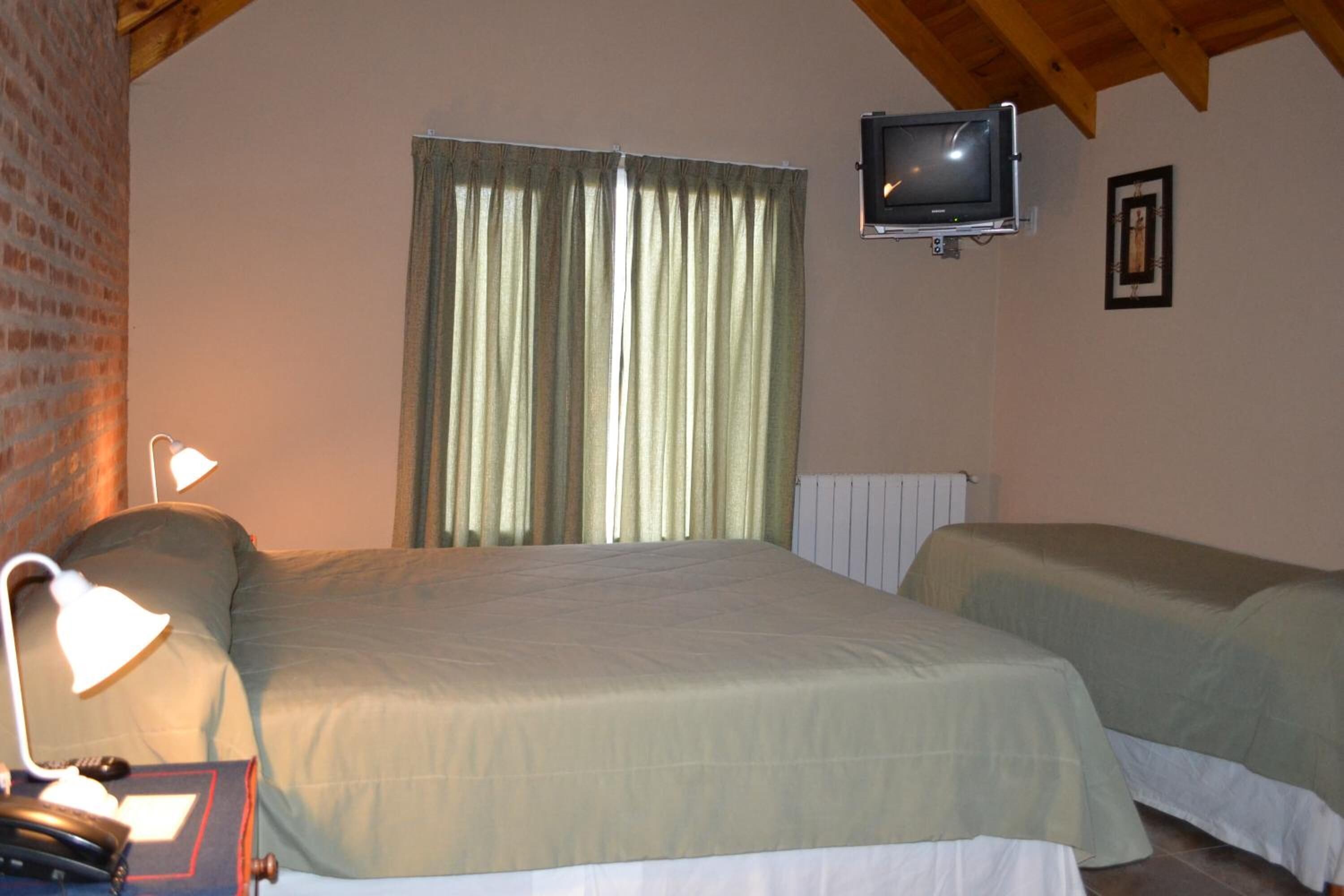 A bedroom with a bed, a desk with a lamp, a television mounted on the wall, and a window with curtains.