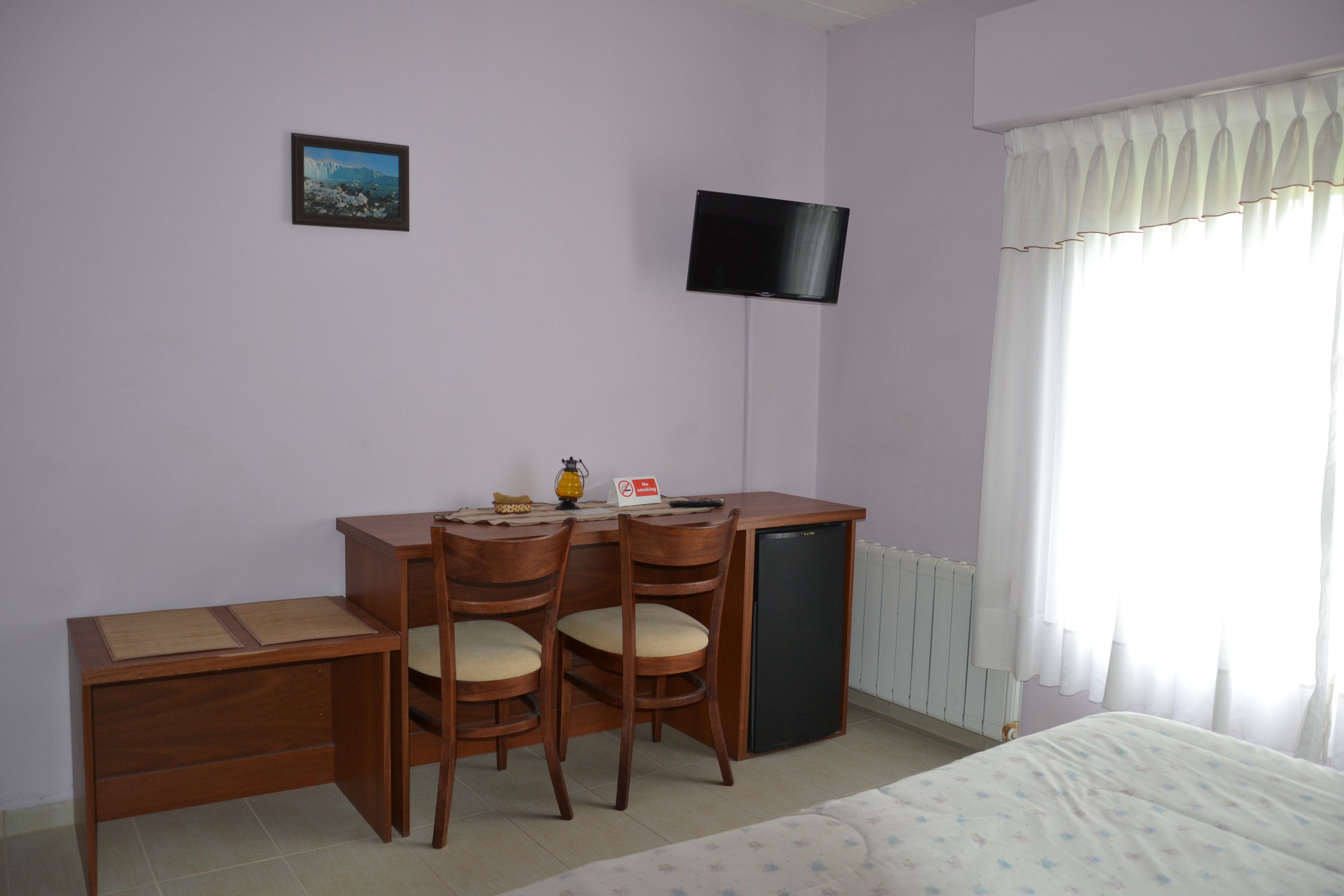 A bedroom with a bed, a desk with two chairs, a television, and a window with curtains.