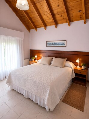 A bedroom with a large bed, wooden ceiling beams, a cityscape picture on the wall, and a bedside table with a lamp. - Alto Verde Hosteria (El Calafate)