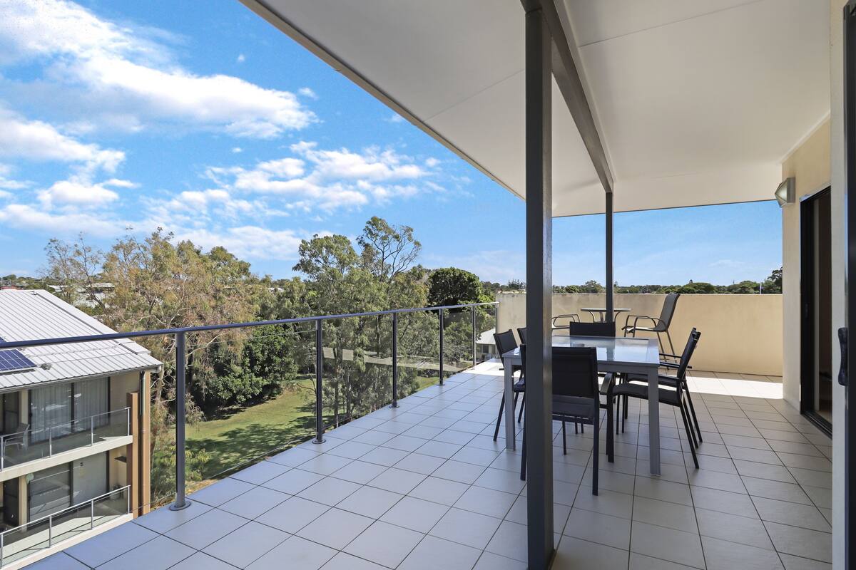 A balcony with a dining table and chairs, a glass railing, and a view of trees and buildings.